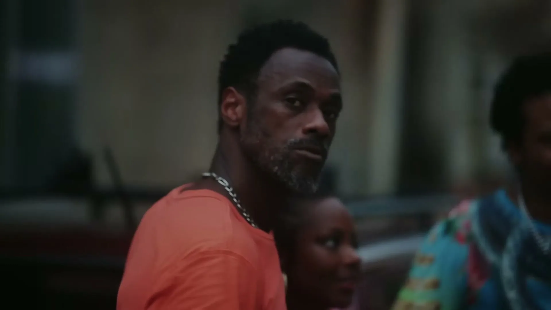 A man with a beard and an orange shirt looks around, surrounded by other people in a lively outdoor setting.