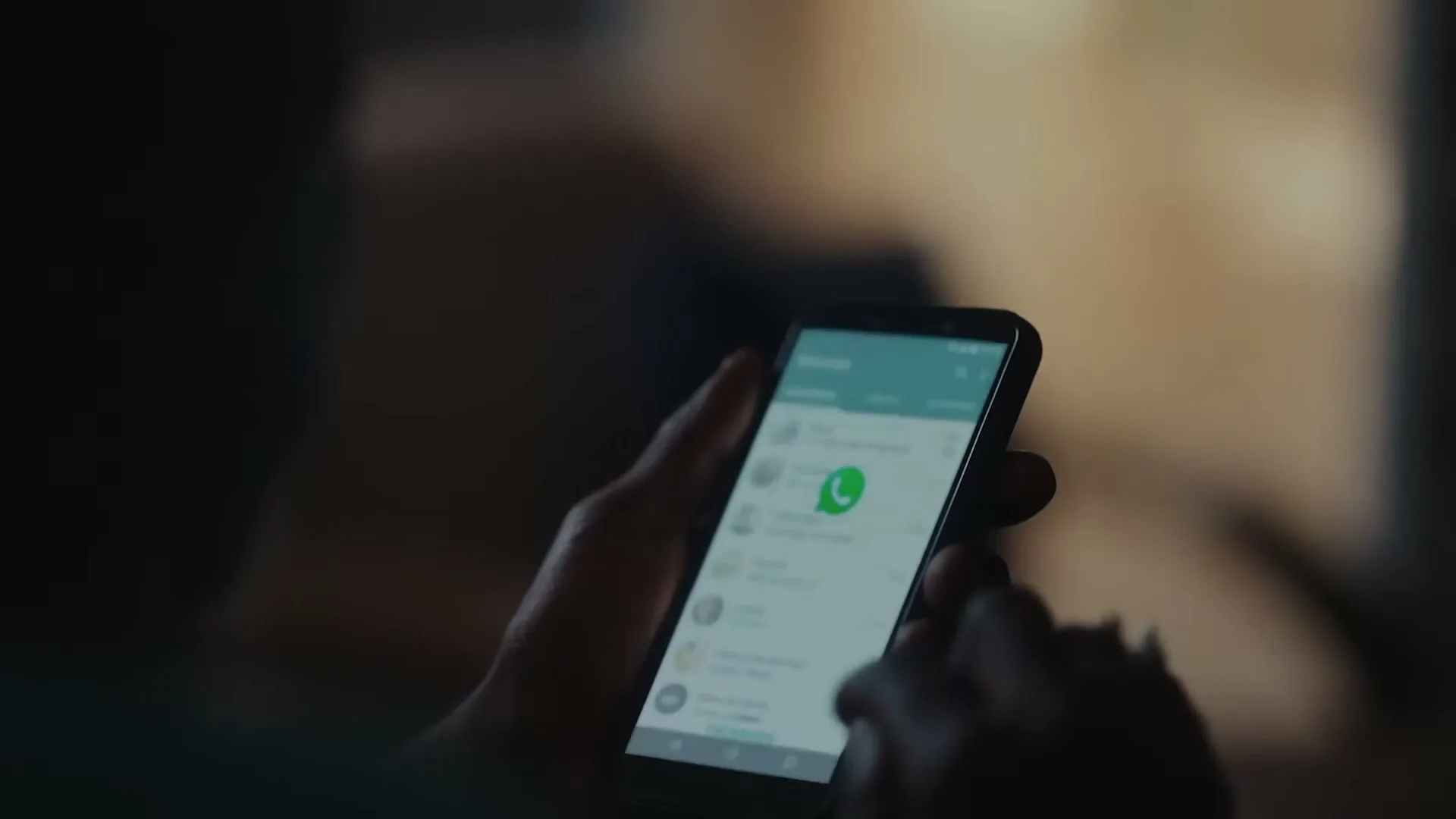 A person's hands are seen in medium closeup, holding a smartphone with the WhatsApp app open, scrolling through contacts in a dimly lit interior.