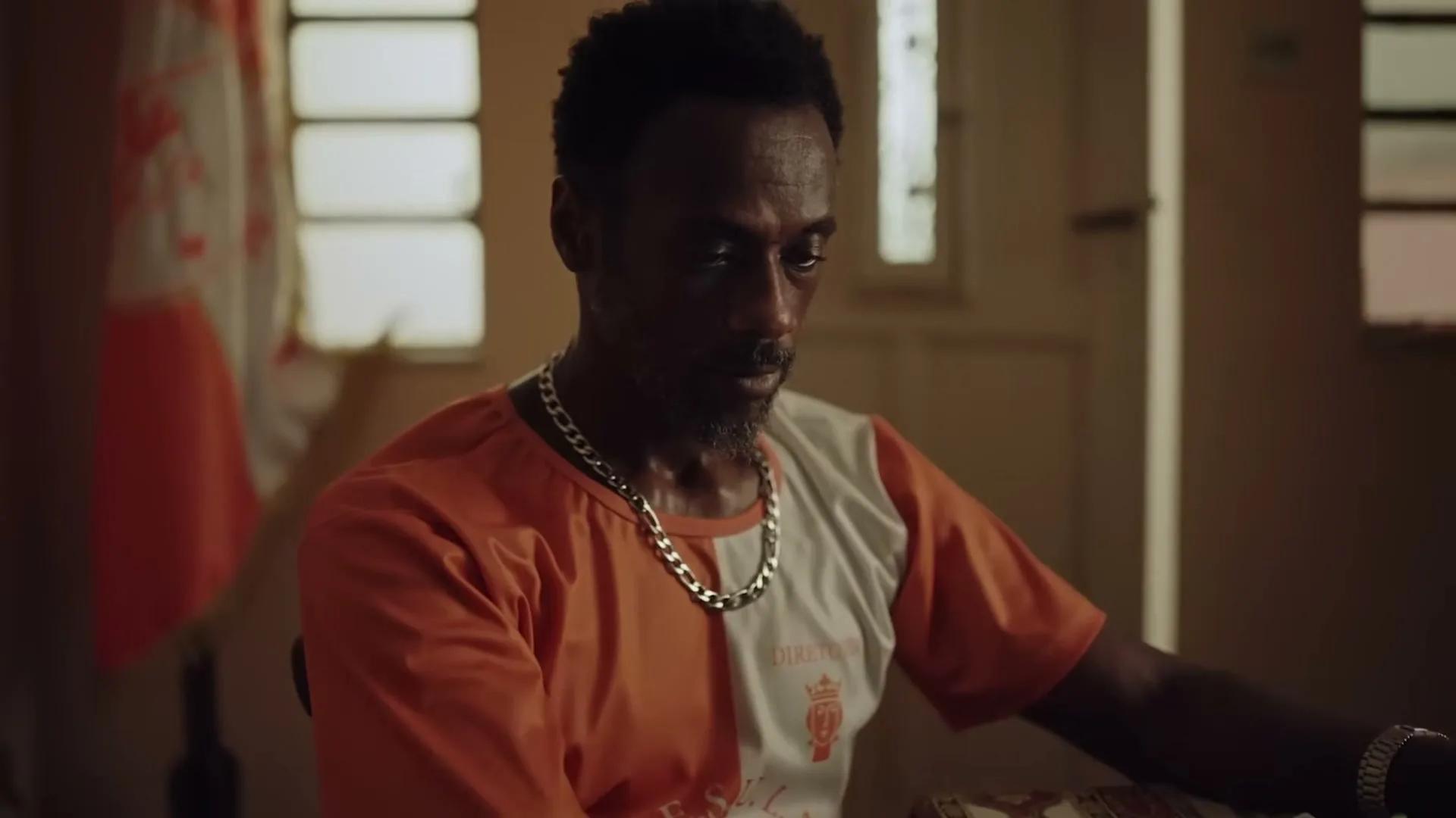 A man with a beard and an orange and white shirt looks down at his phone while sitting at a table with food.