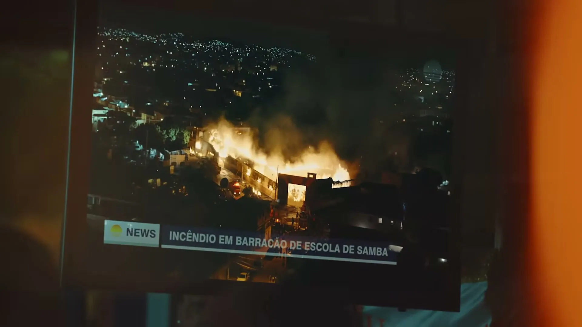 A news report on a TV screen shows an aerial view of a large fire burning intensely in a city at night.