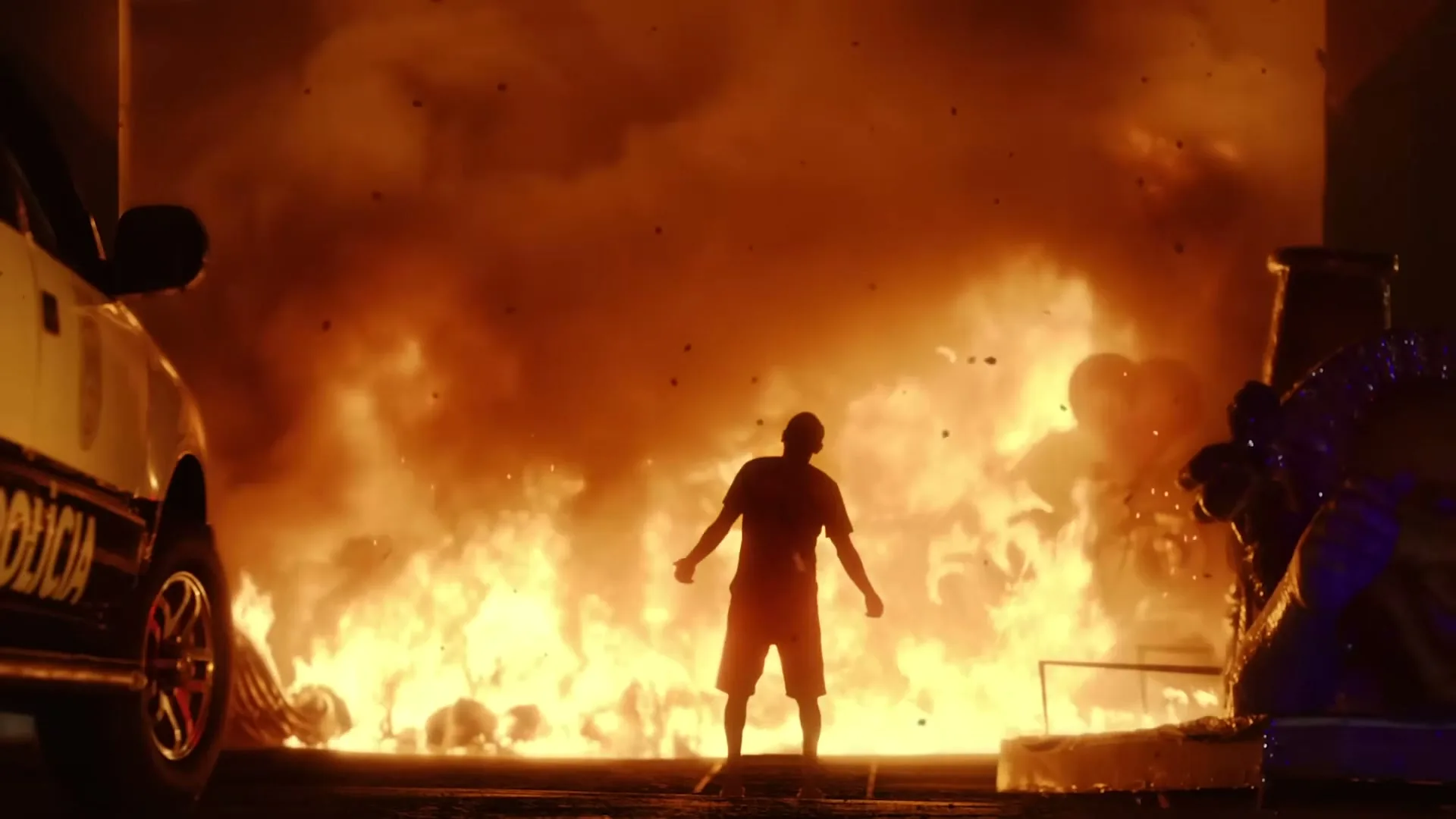 A man stands silhouetted against a massive fire, with a police car partially visible on the left and a carnival float on the right, at night.