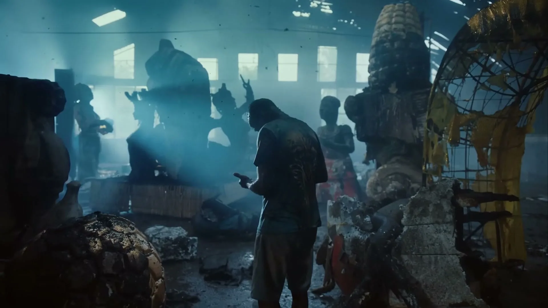 A man stands in a smoky, dimly lit warehouse, looking at his phone, surrounded by large, partially destroyed carnival floats and other people.