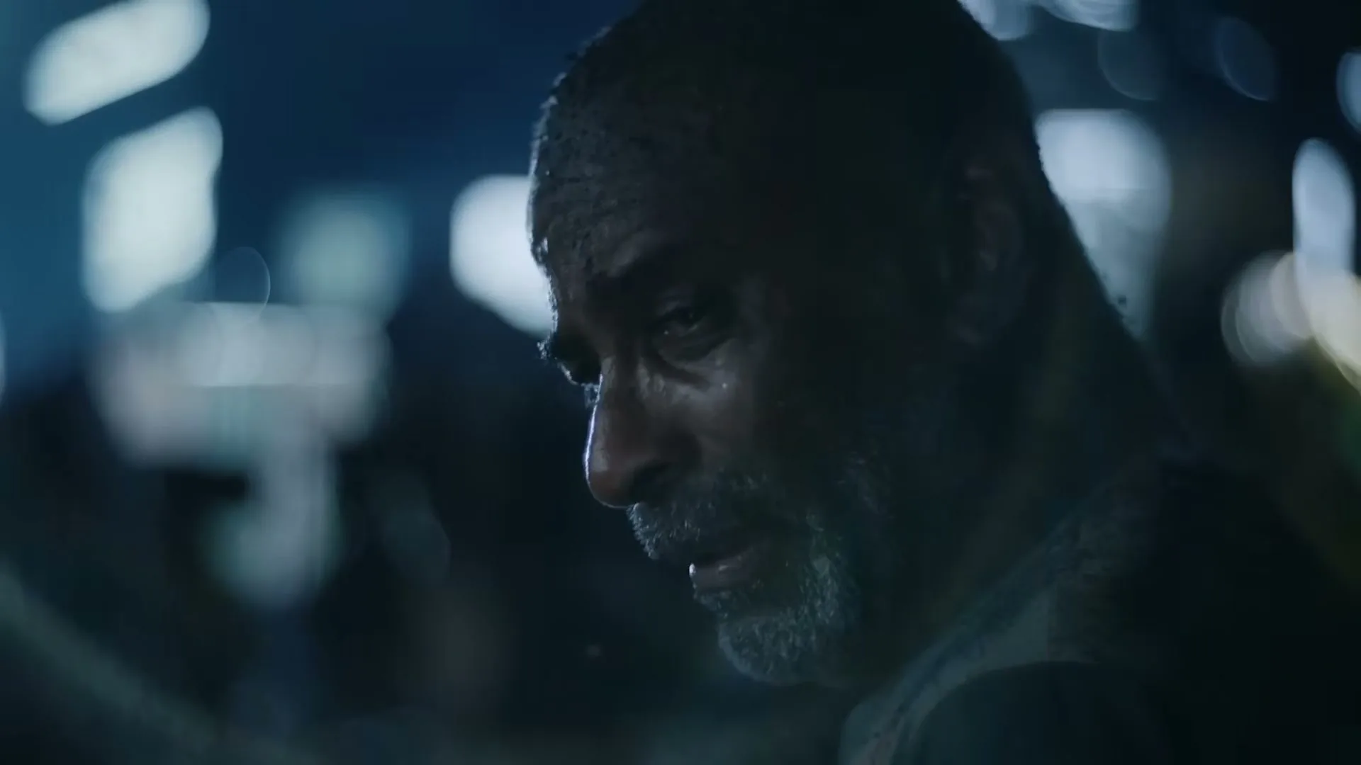 A man with a beard and wet hair looks up with a sad expression, illuminated by blue light in a dark, blurry background.