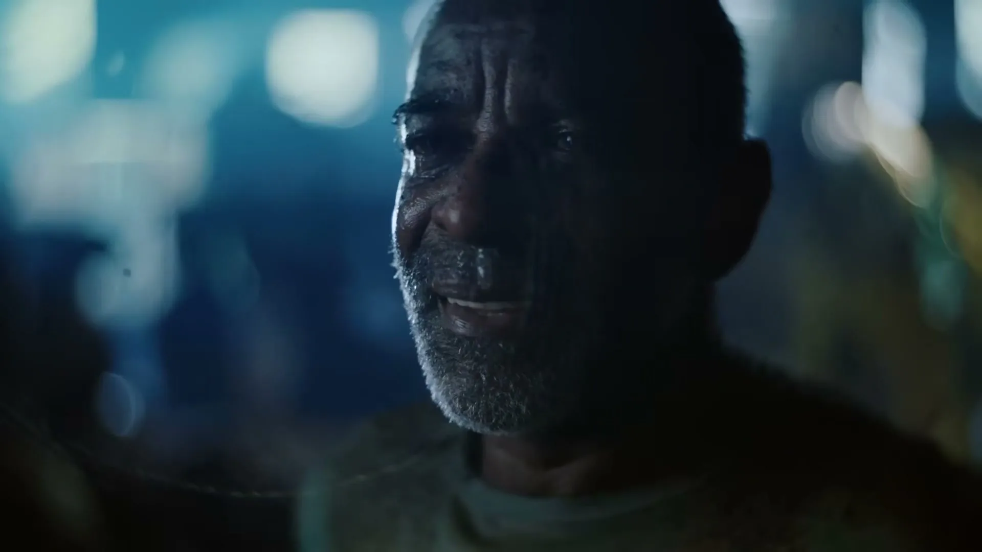 A man with a grey beard and wet face looks up with a slight smile, illuminated by blue light in a dark, blurry background.