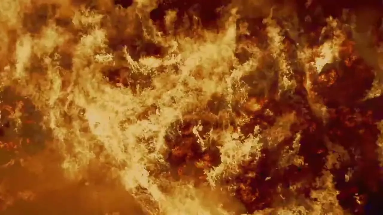An extreme wide shot shows a large area of land engulfed in bright orange and yellow flames, with smoke rising.