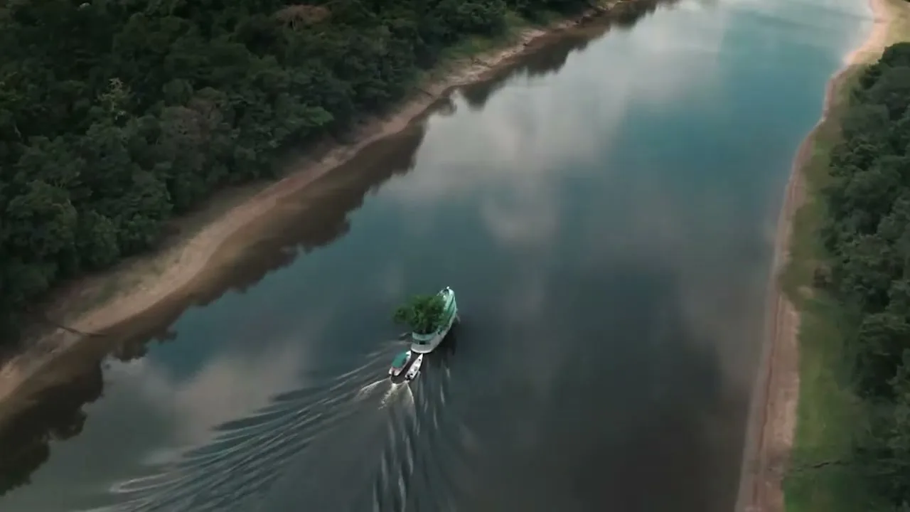 An aerial shot shows a boat with a tree on it, pulling two smaller boats, traveling down a wide river surrounded by dense green jungle.