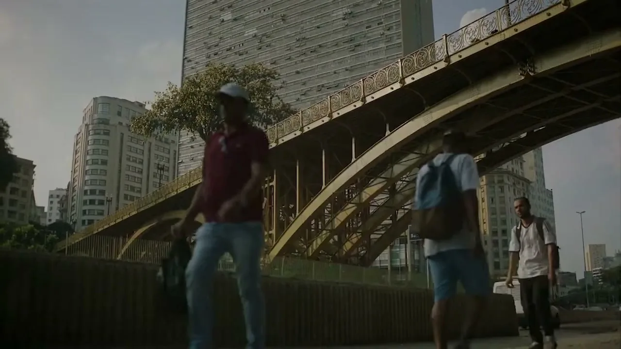A wide shot shows a tree growing on a bridge over a busy street, with people walking by and tall buildings in the background.