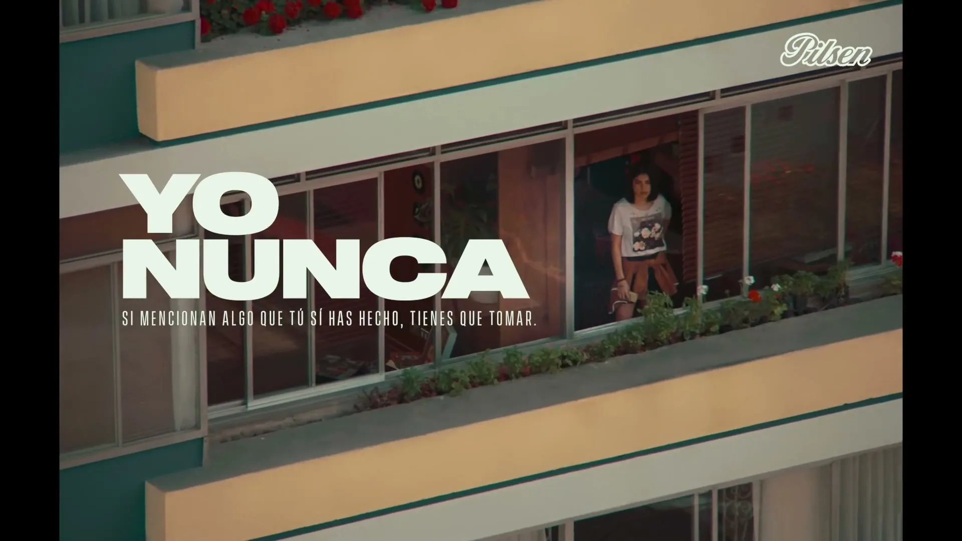 A young woman in a white t-shirt and brown pants stands on a balcony of a teal and yellow apartment building, looking out, with text overlay.