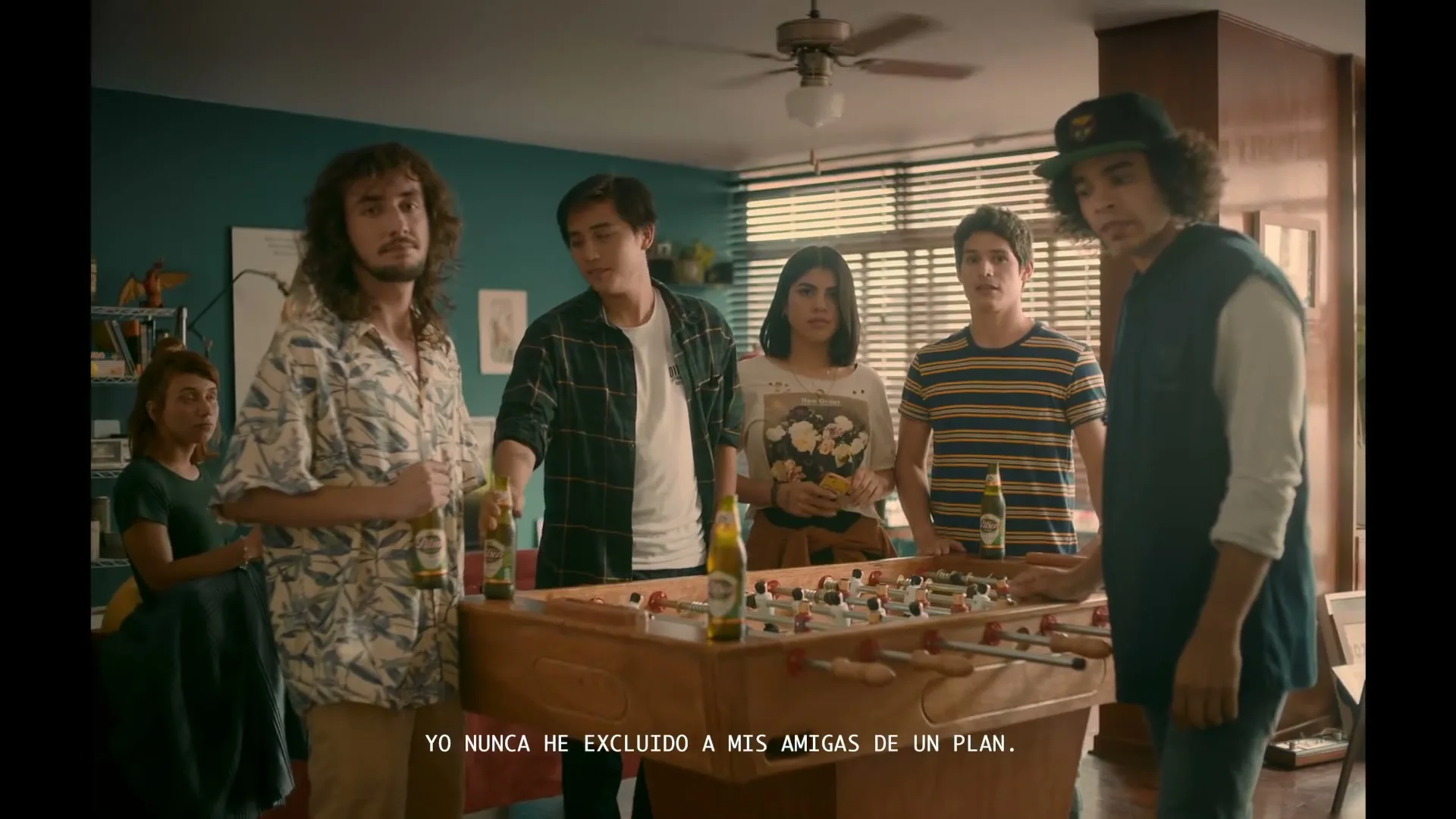 A group of young adults, including three men and two women, are gathered around a foosball table in a living room, playing and drinking beer.