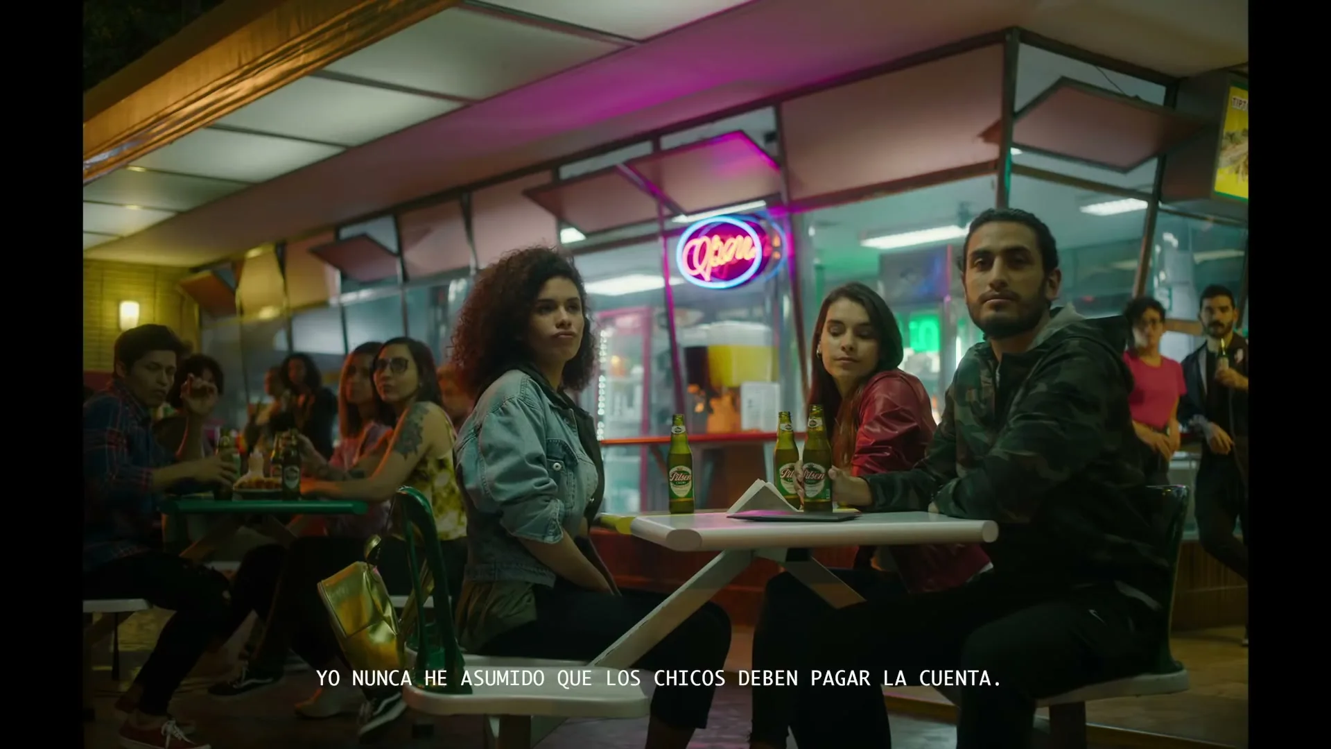 A diverse group of young adults sits at outdoor tables at night, drinking beer and listening intently to a man speaking.
