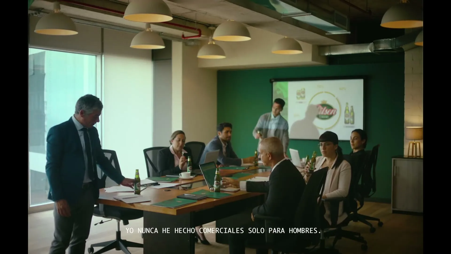 A diverse group of business people in a modern conference room, some standing and some seated, all drinking from green bottles of Pilsen Callao beer.