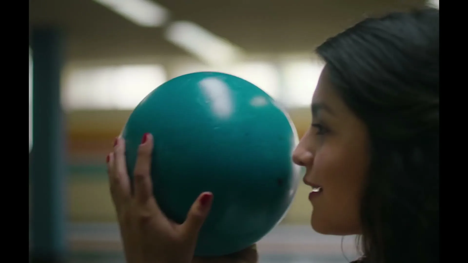 A young woman with dark hair and red nail polish holds a teal bowling ball, then turns her head to the left in a bowling alley.