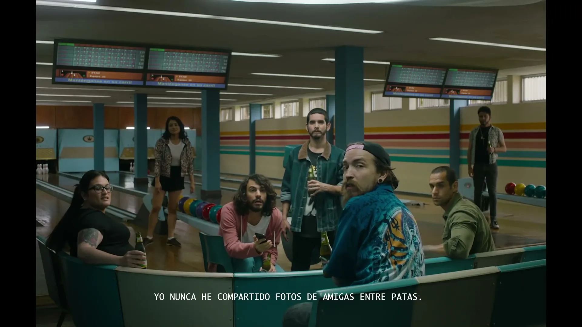 A group of friends, including men with beards and a woman in glasses, are sitting and standing in a bowling alley, drinking beer and looking at a phone.