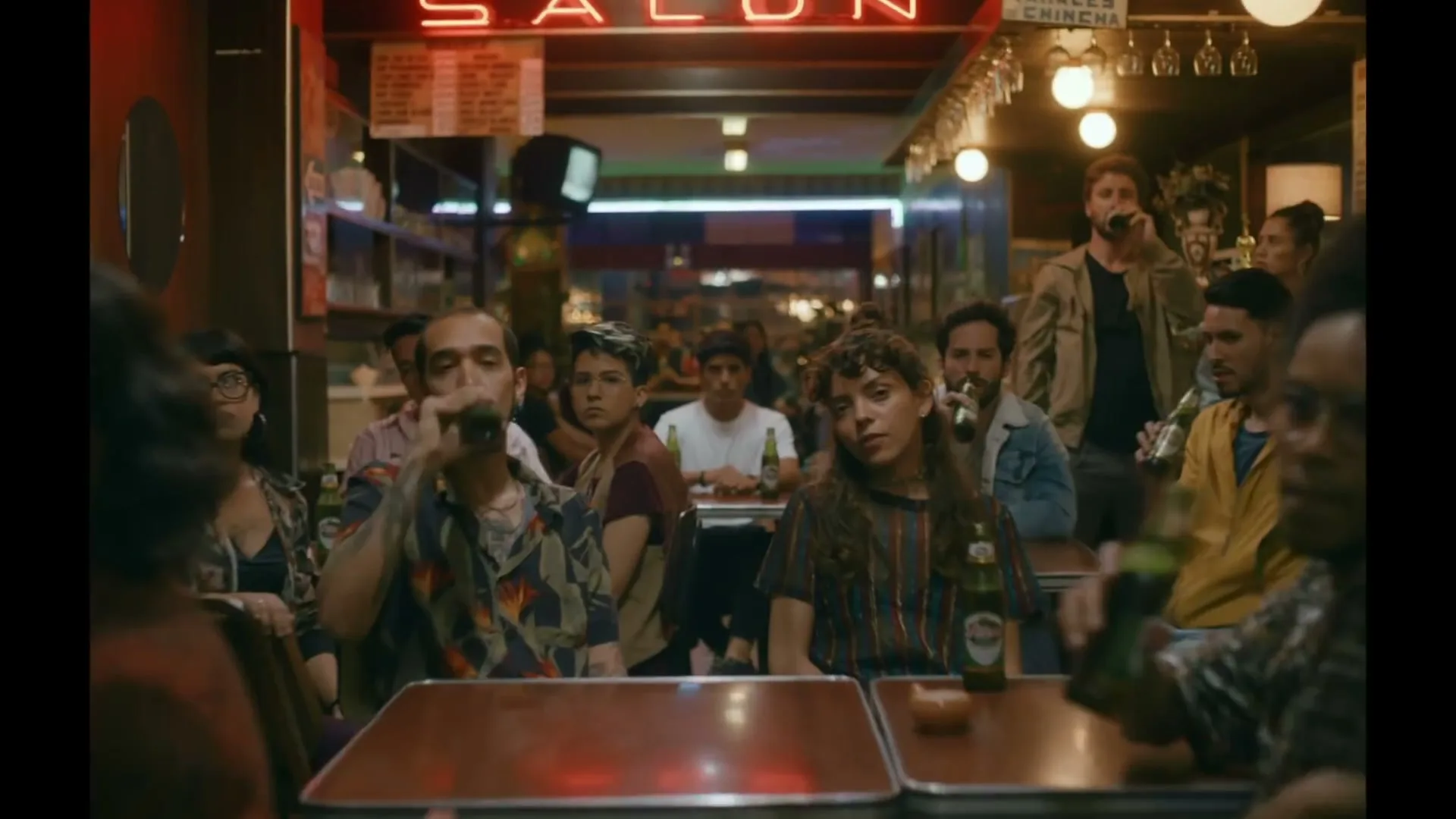 A diverse group of young adults sits at tables in a dimly lit bar, some drinking beer, as the camera slowly pushes in on a couple in the foreground.