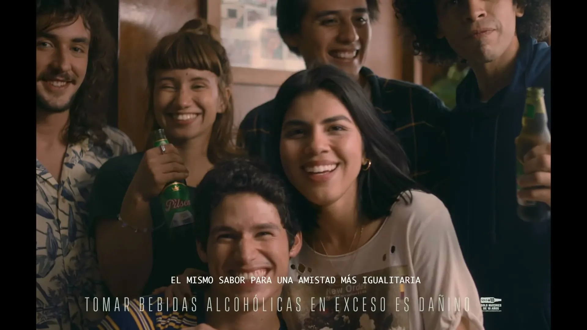 A diverse group of young adults, smiling and holding beer bottles, pose for a photo in a warmly lit interior setting.