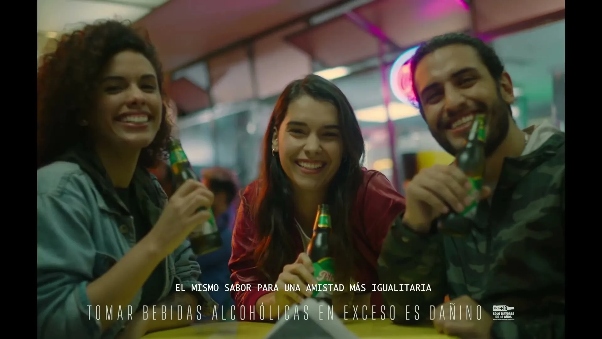 Three young adults, two women and one man, are sitting at a table, holding green beer bottles and smiling happily at the camera in an outdoor setting at night.