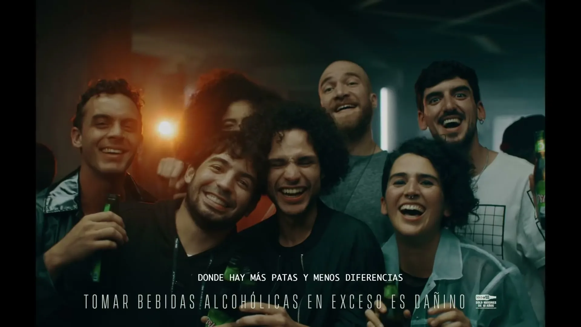 A diverse group of young adults, holding beer bottles, laugh and smile at the camera in a dimly lit, industrial-style interior.