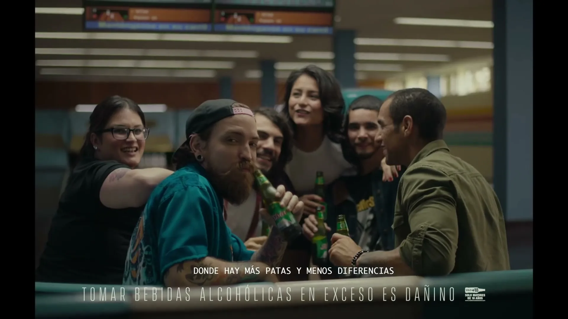 A diverse group of friends, including a bearded man in a blue shirt, are smiling and drinking beer together at a bar in an interior setting.