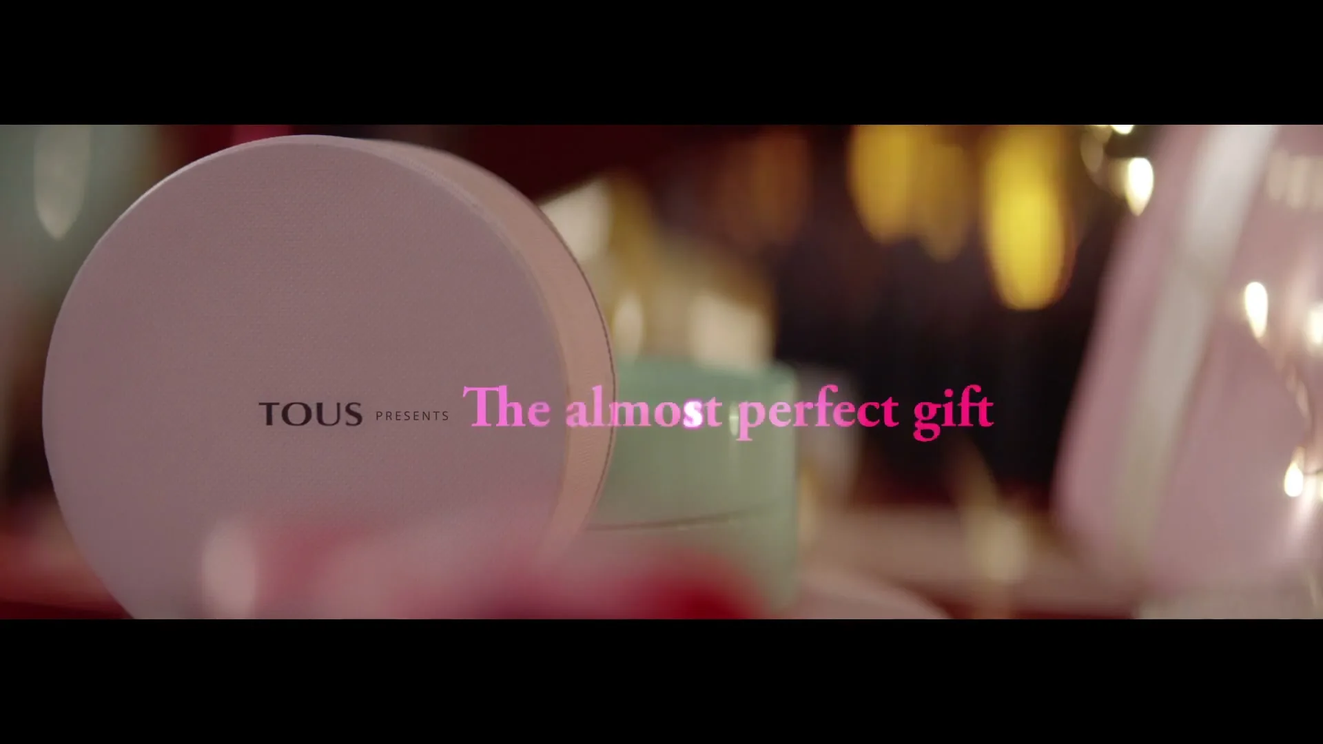 A light pink round gift box with "TOUS PRESENTS" and "The almost perfect gift" text on it, surrounded by blurred lights.