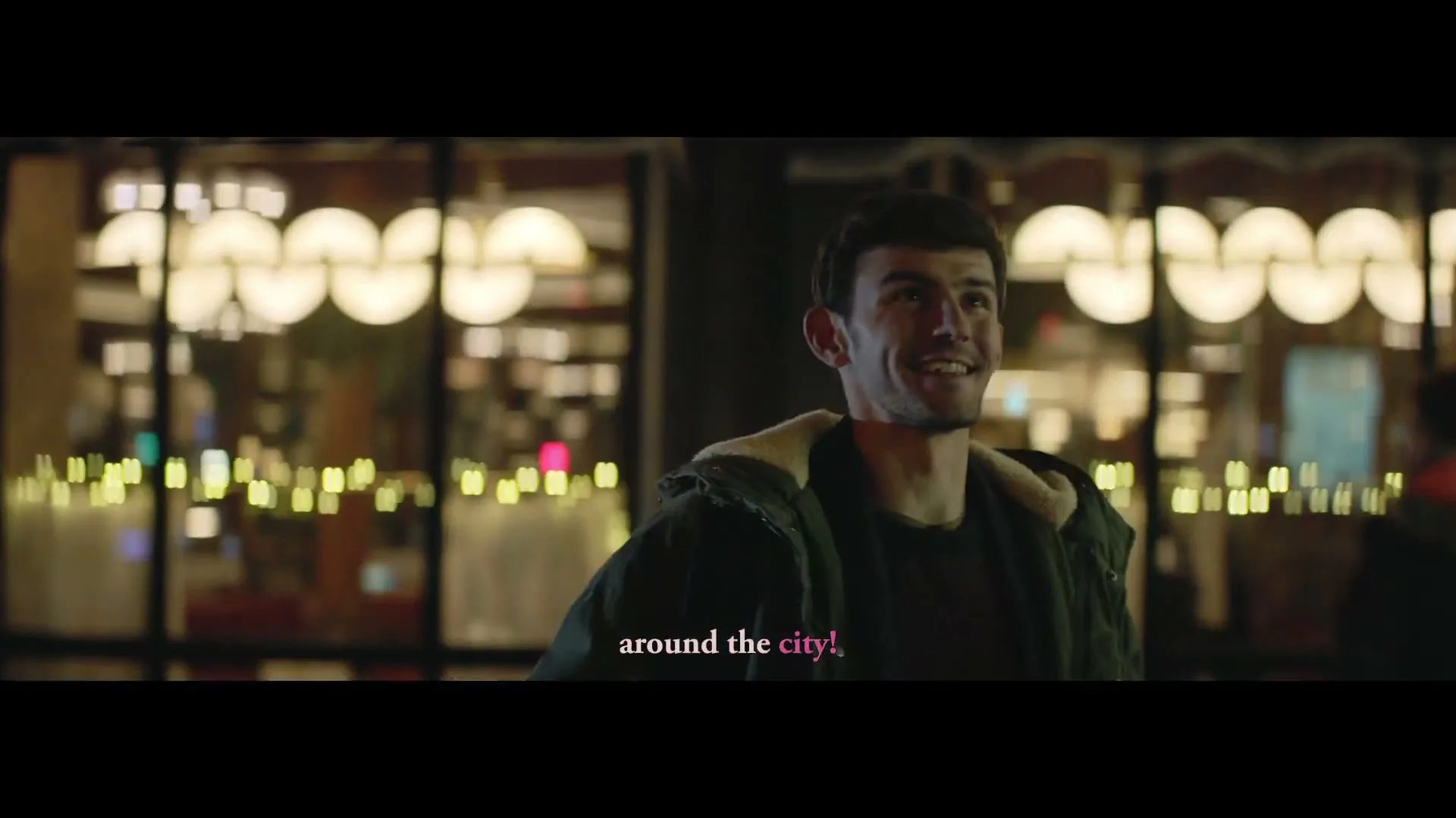 A young man in a winter coat smiles and looks up, standing on a city street at night with blurred lights in the background.