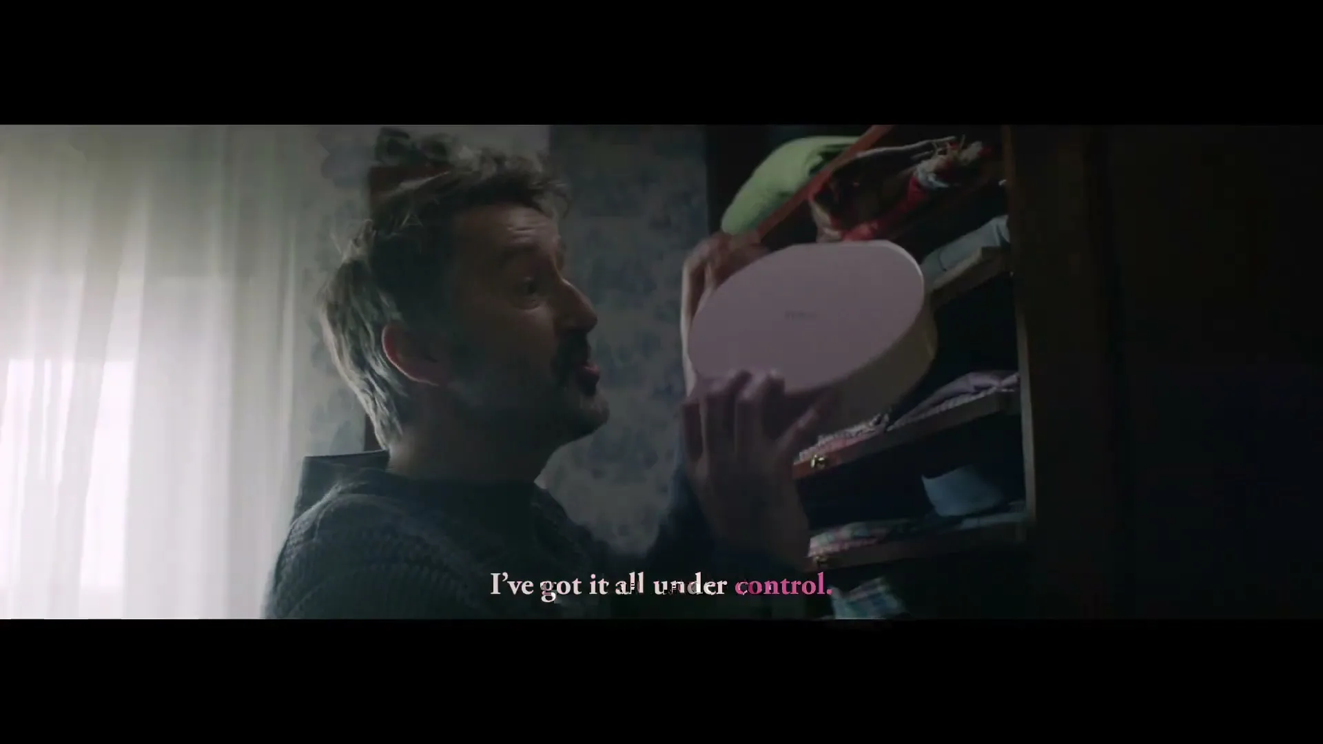 A man in his 40s, with a beard and messy hair, pulls a pink box from a closet shelf in a bedroom.