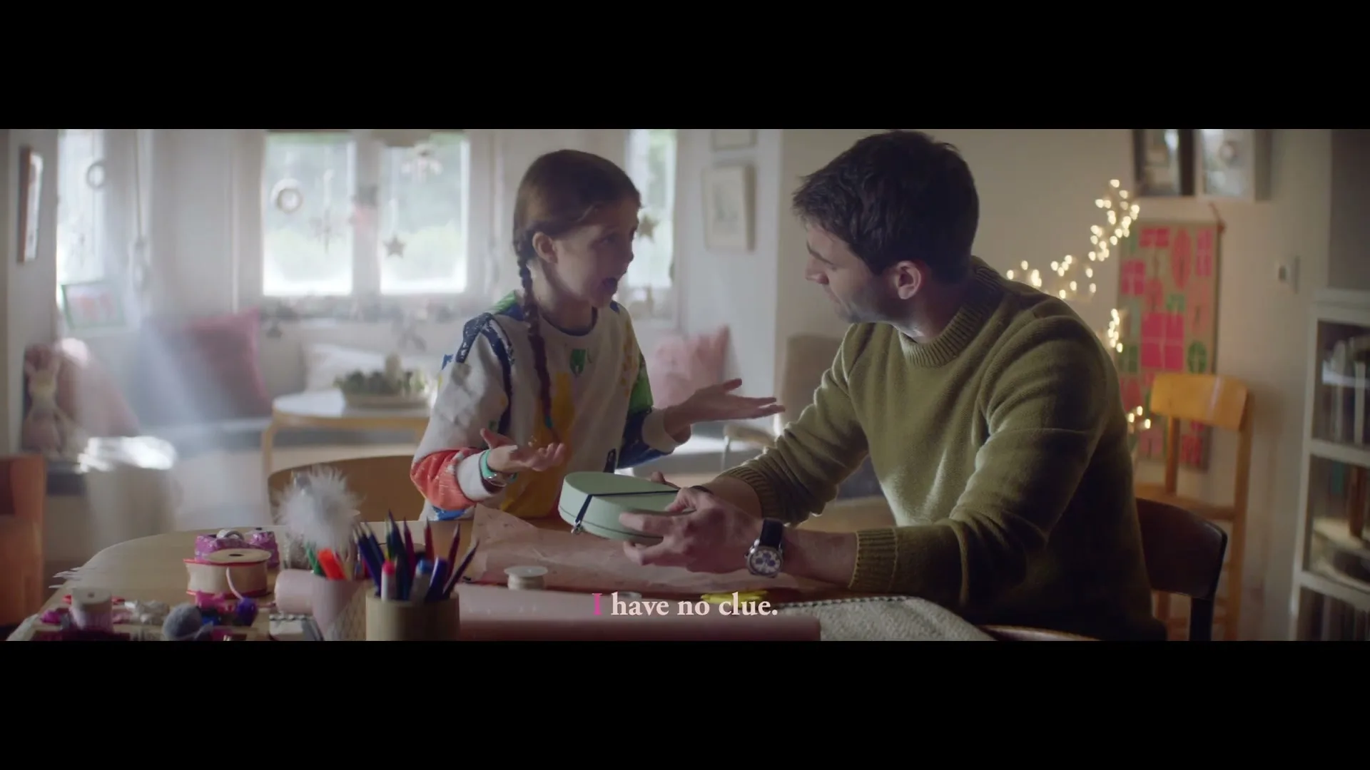 A young girl and a man sit at a table, wrapping a gift in a cozy, decorated room, with sunlight streaming through a window.