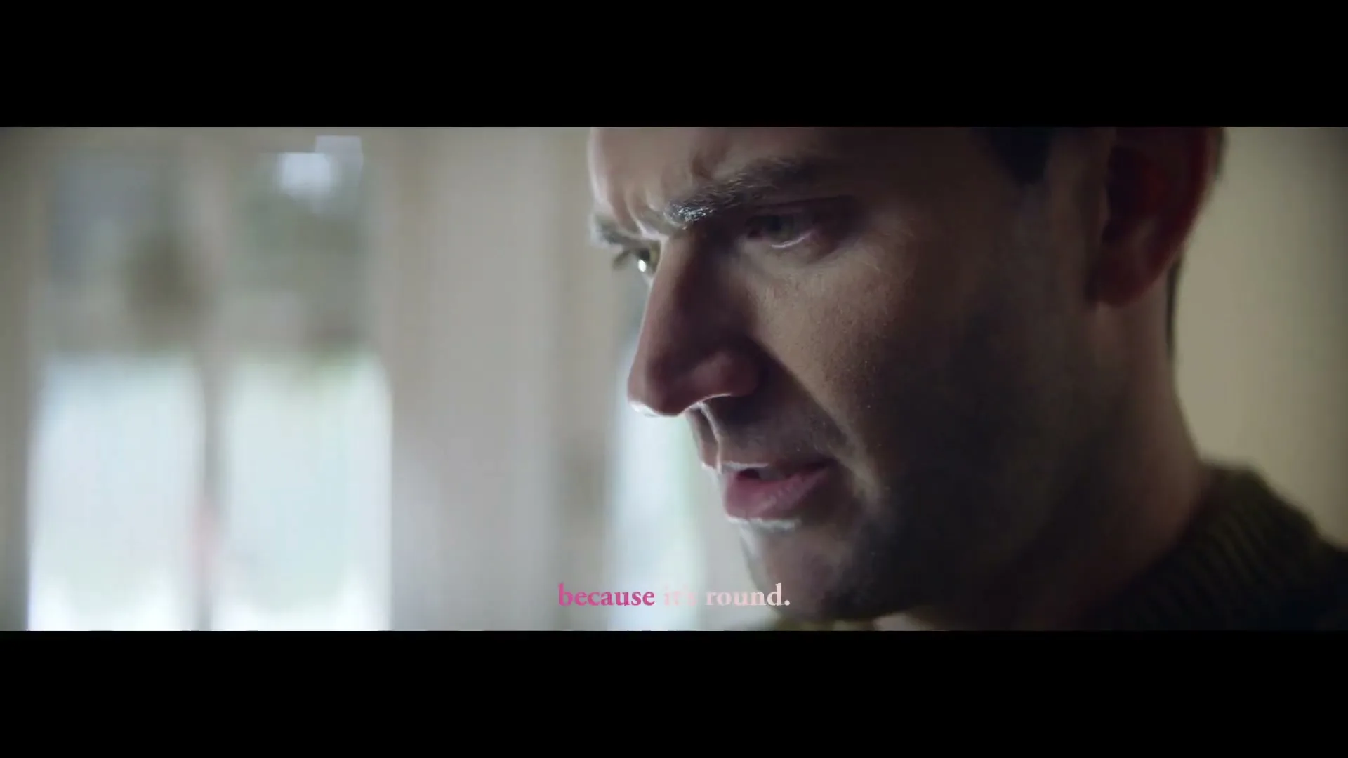 A man in his 30s, wearing a green sweater, looks down with a concerned expression in an interior setting.