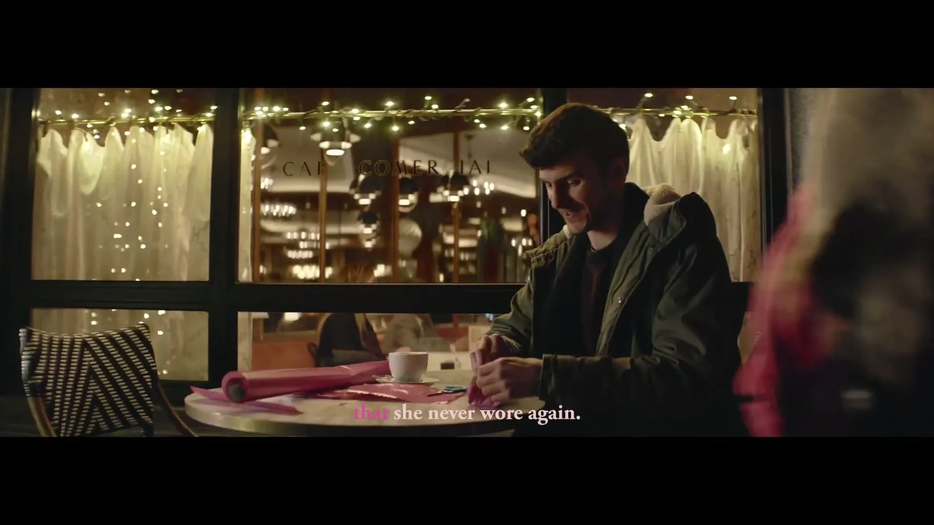 A young man with dark hair and a green jacket sits at a cafe table, smiling as he wraps a small gift with pink paper, with string lights visible through the window.