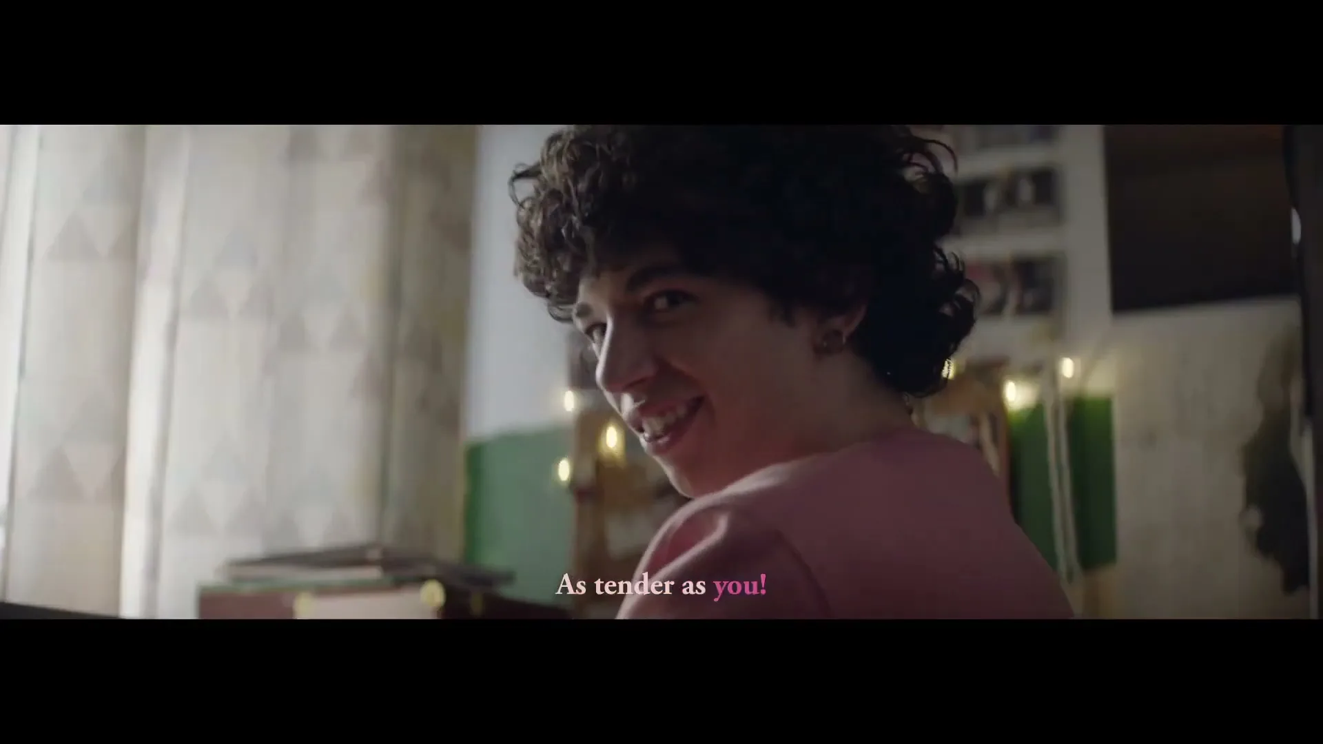 A young man with curly hair, wearing a pink shirt, turns his head to look back at the camera and smiles mischievously in a cozy room.