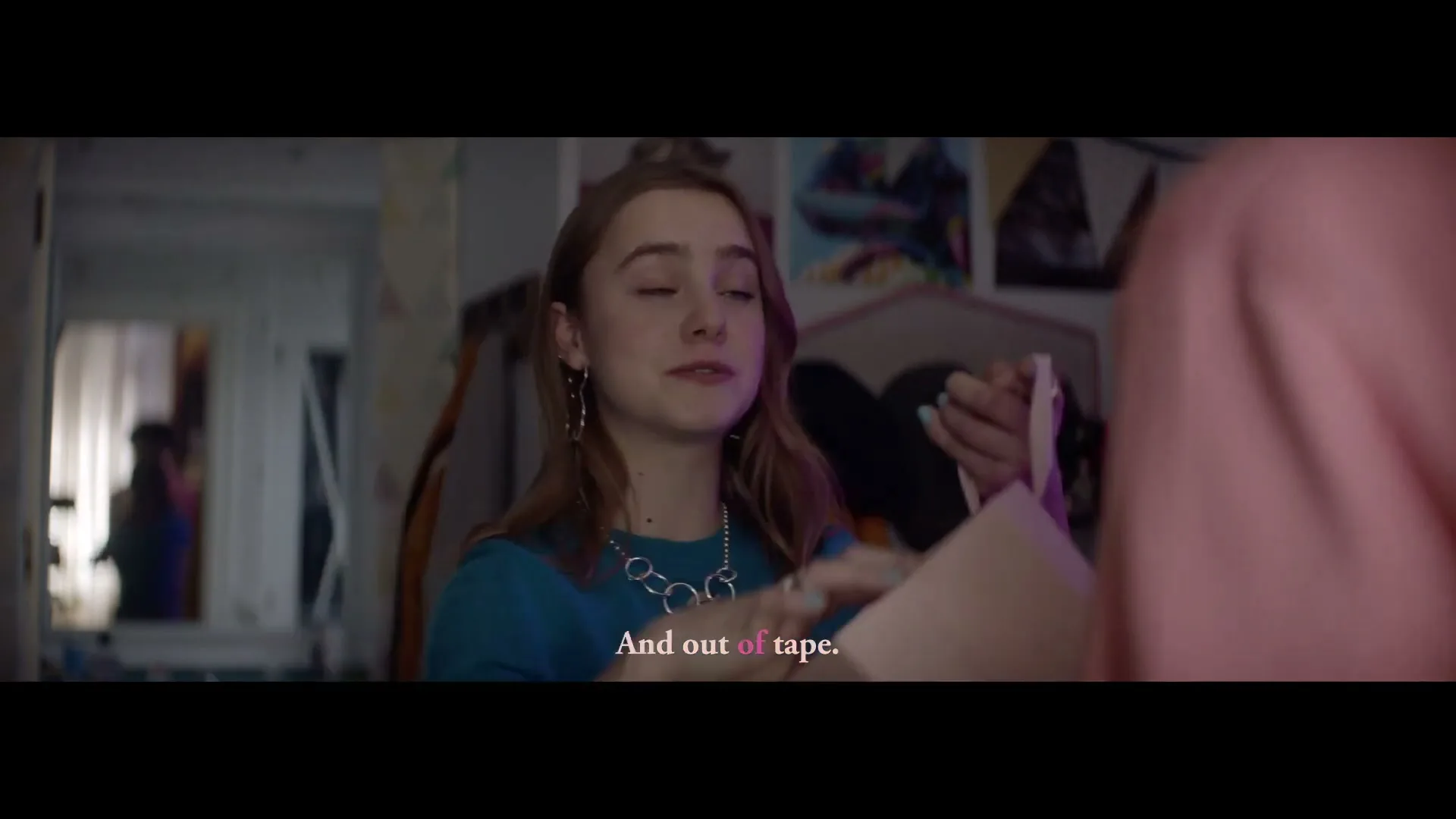A young woman with long brown hair and a blue top talks to someone off-screen, holding a pink object in her hands in a bedroom.