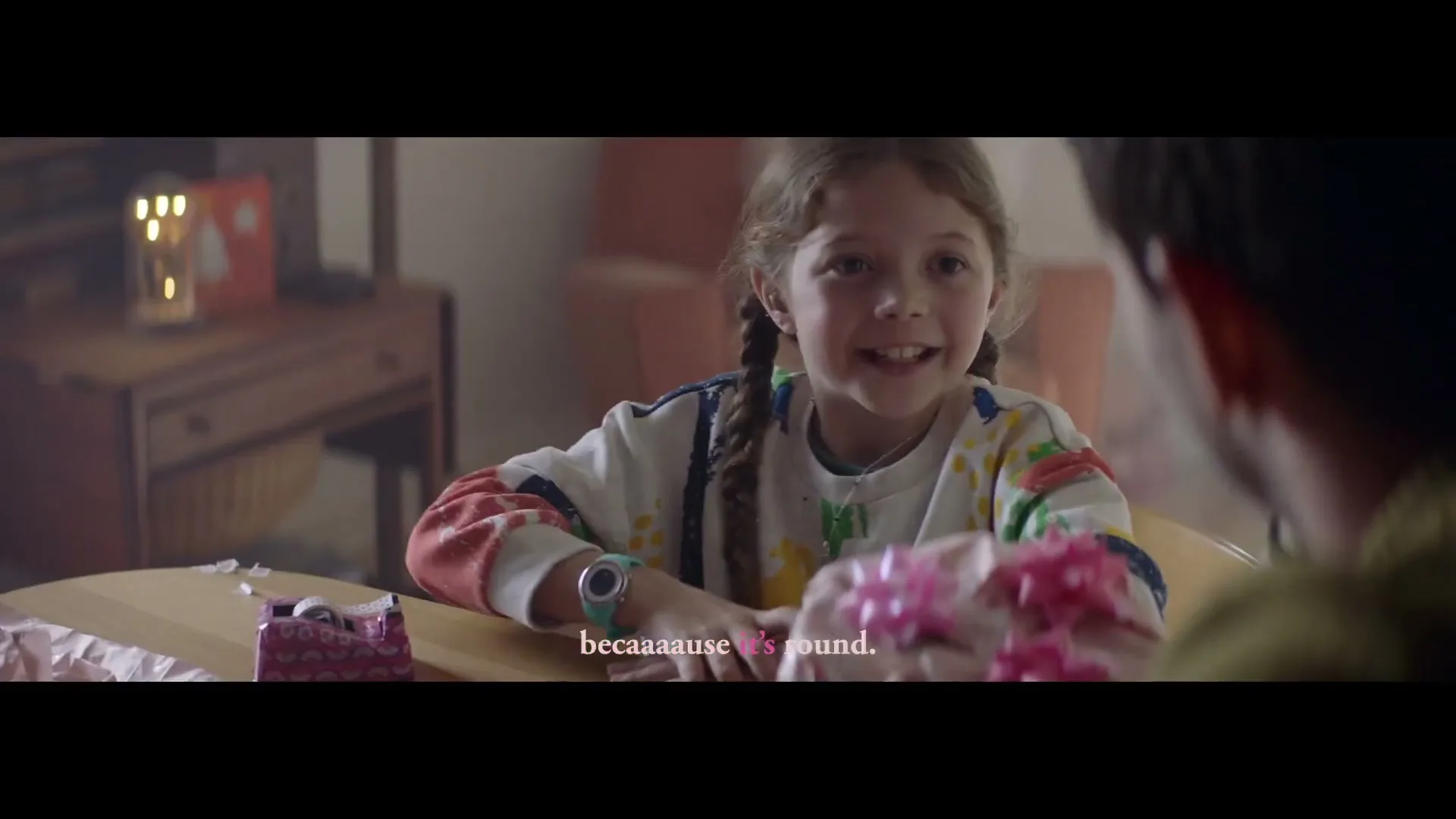 A young girl with braided hair, wearing a colorful sweatshirt, smiles happily as she hands a wrapped gift to an unseen person across a wooden table in a cozy room.