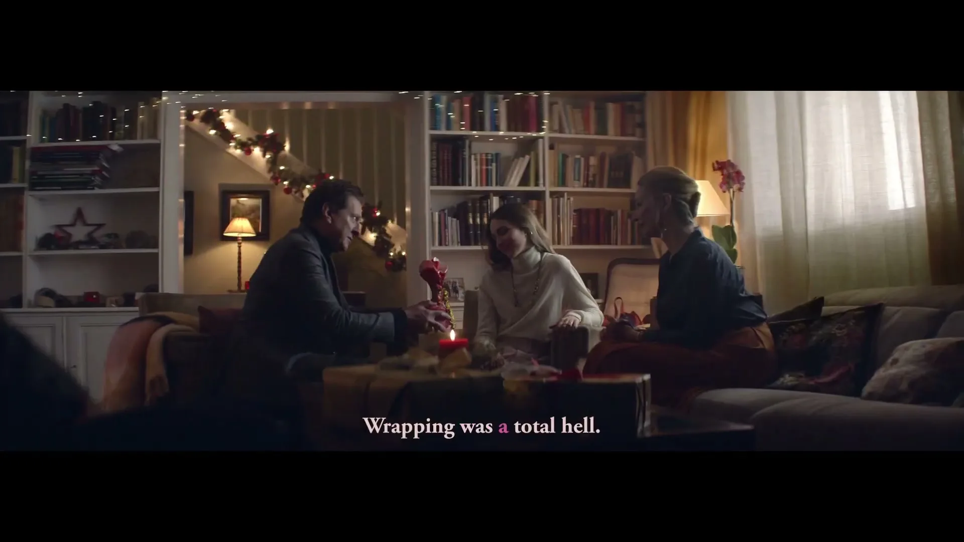 A man in a dark suit hands a small gift to a woman in a white sweater, while another woman watches, in a cozy living room decorated for Christmas.