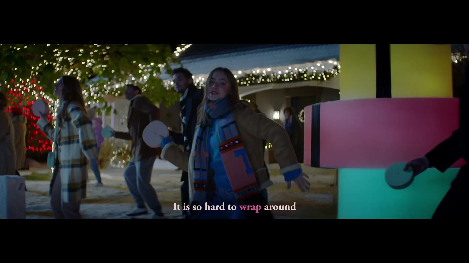 A young woman in a winter coat and scarf dances with a white disc in her hand, surrounded by other people holding discs, in a festive outdoor setting at night.