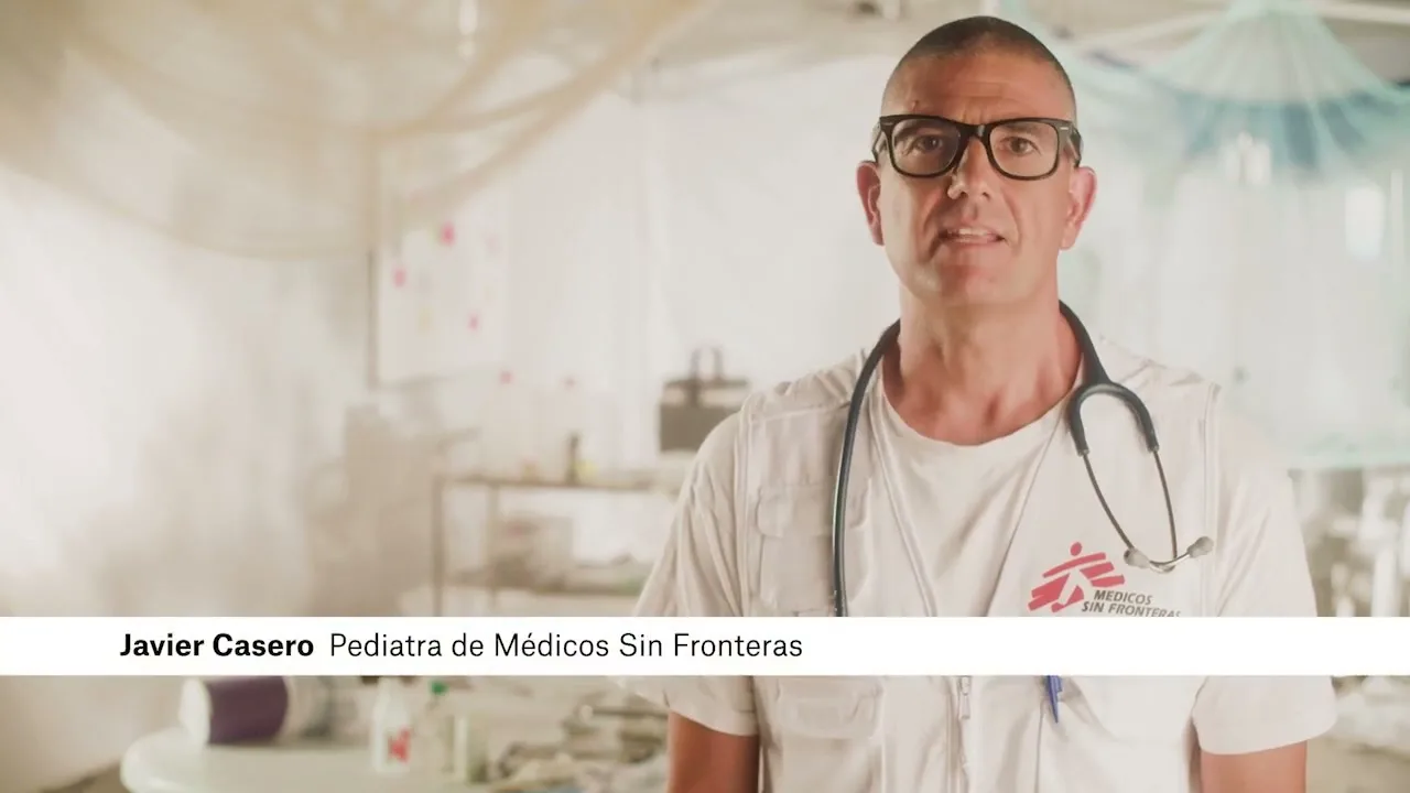 A male pediatrician wearing glasses and a stethoscope speaks directly to the camera in a medical tent.