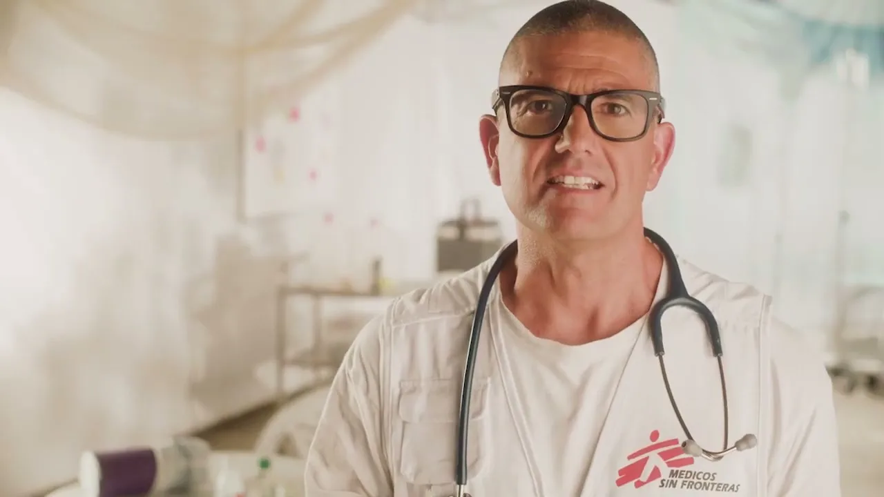 A male doctor with glasses and a stethoscope around his neck speaks directly to the camera in a brightly lit medical tent.