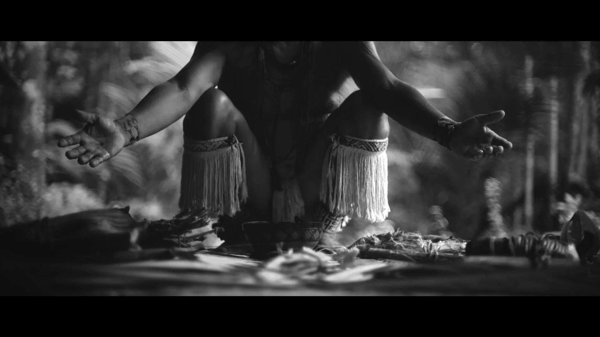 A person with tribal tattoos and traditional attire crouches with open hands over ritualistic items, surrounded by smoke in a forest setting.