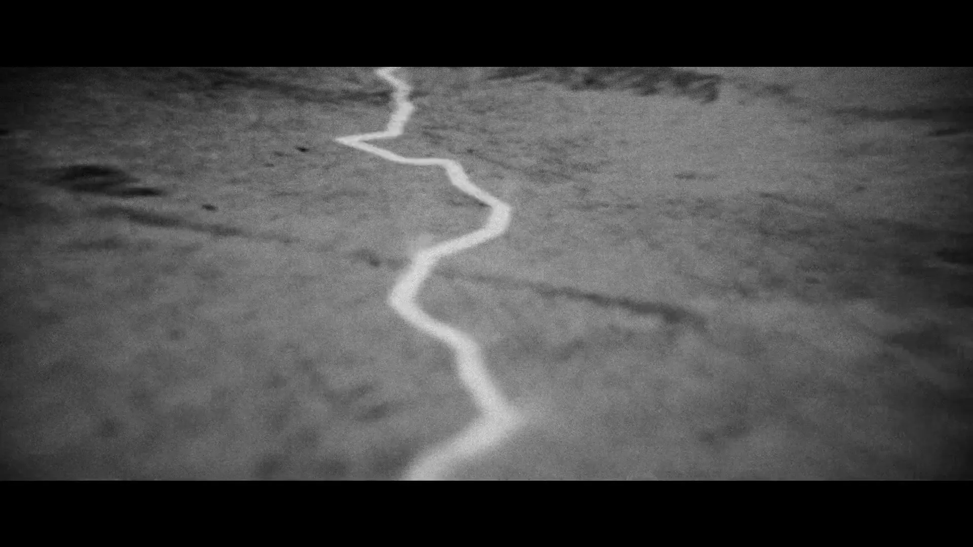 A black and white, grainy, high-angle shot shows a winding, glowing line moving across a textured, dark surface, resembling a river or crack.