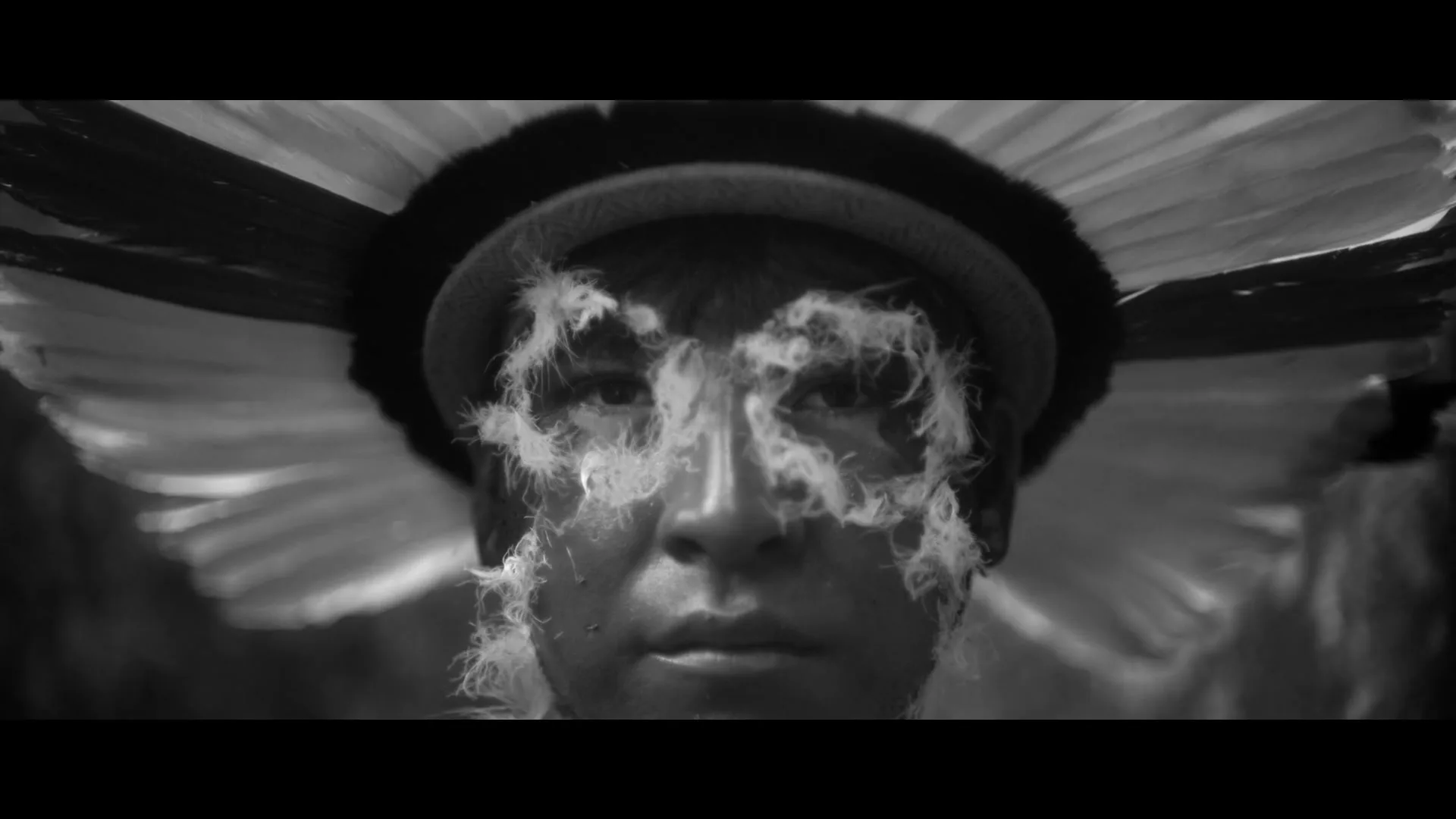 A close-up of an indigenous person's face with feathers around their eyes, transitioning to a medium shot of three indigenous people in a river with a stilt house in the background.