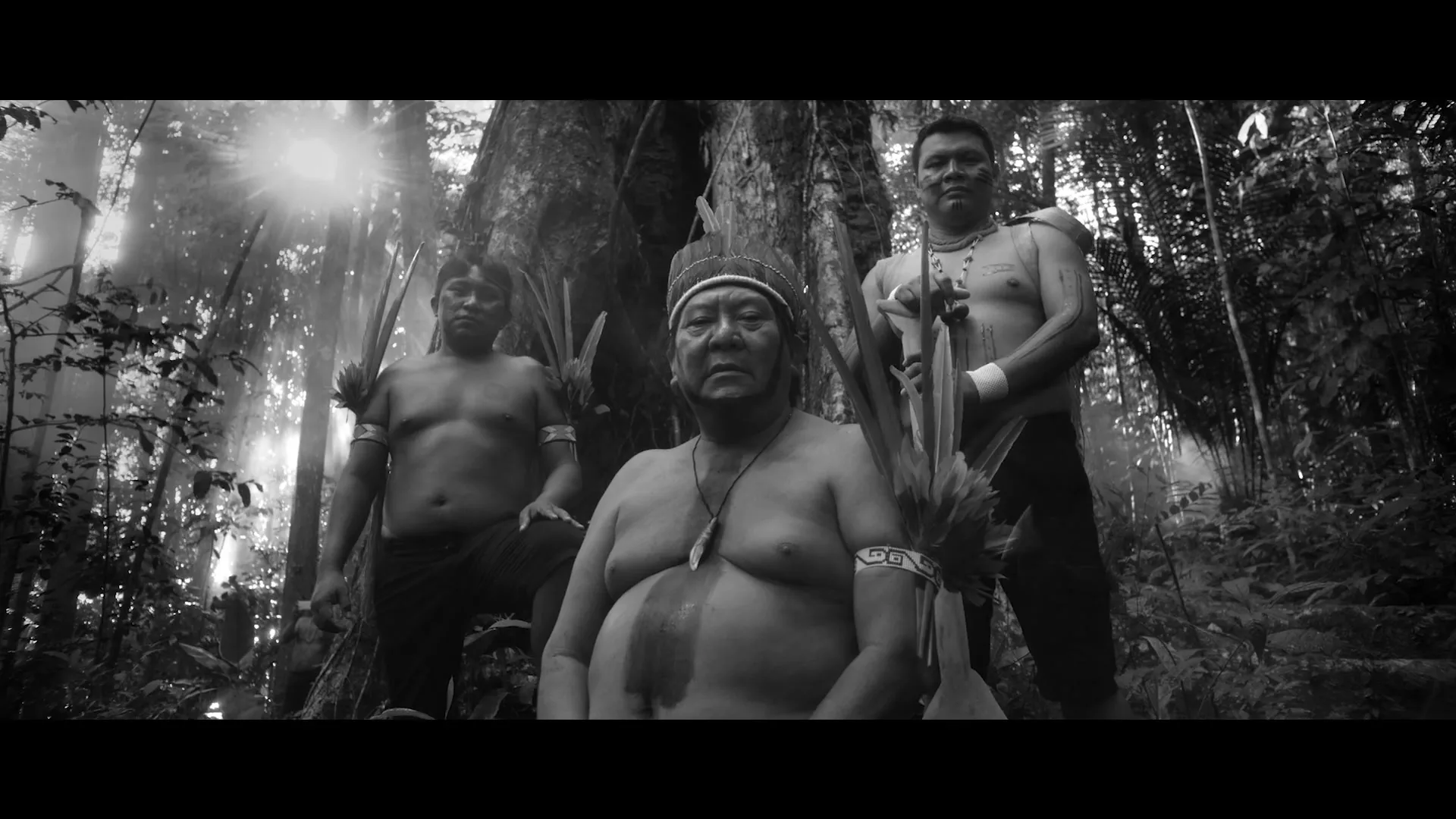 A black and white shot shows a group of indigenous people standing in a dense forest, looking directly at the camera with serious expressions.