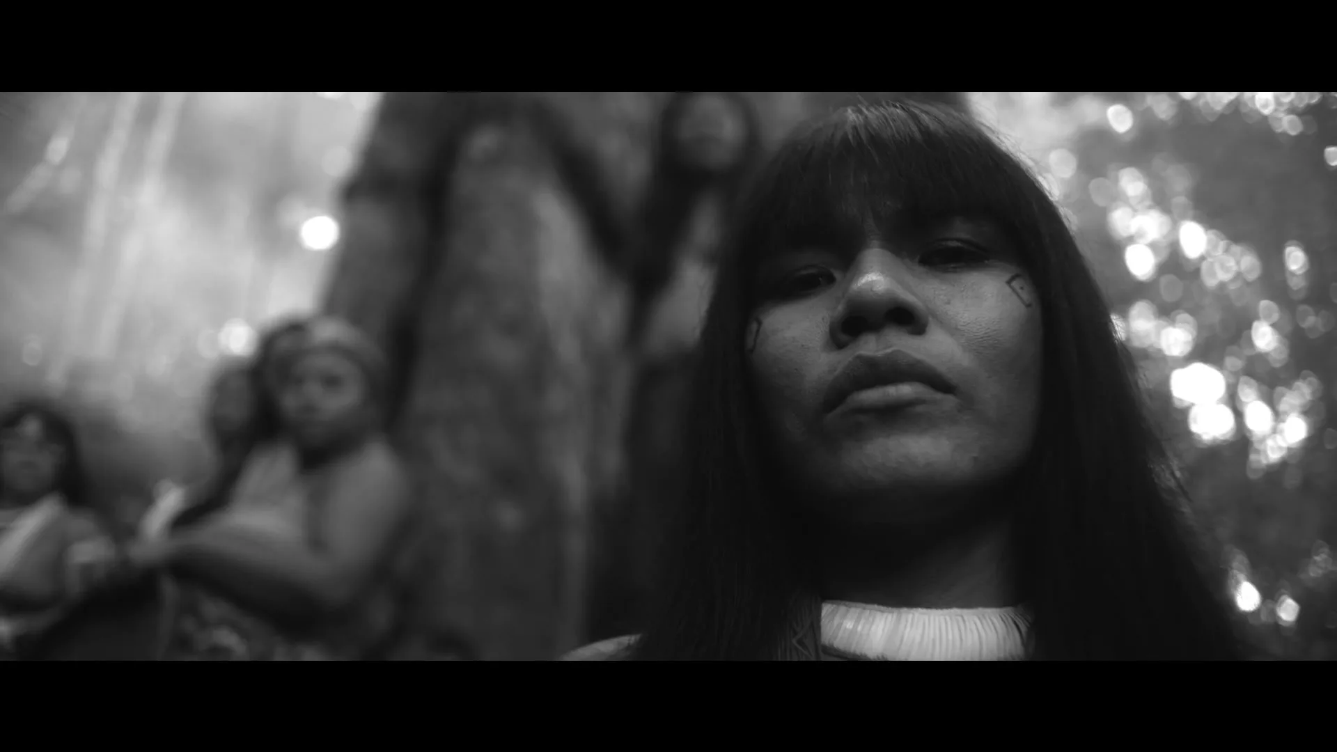 A young Indigenous woman with long dark hair and facial markings looks directly at the camera, with other people blurred in the background of a forest.