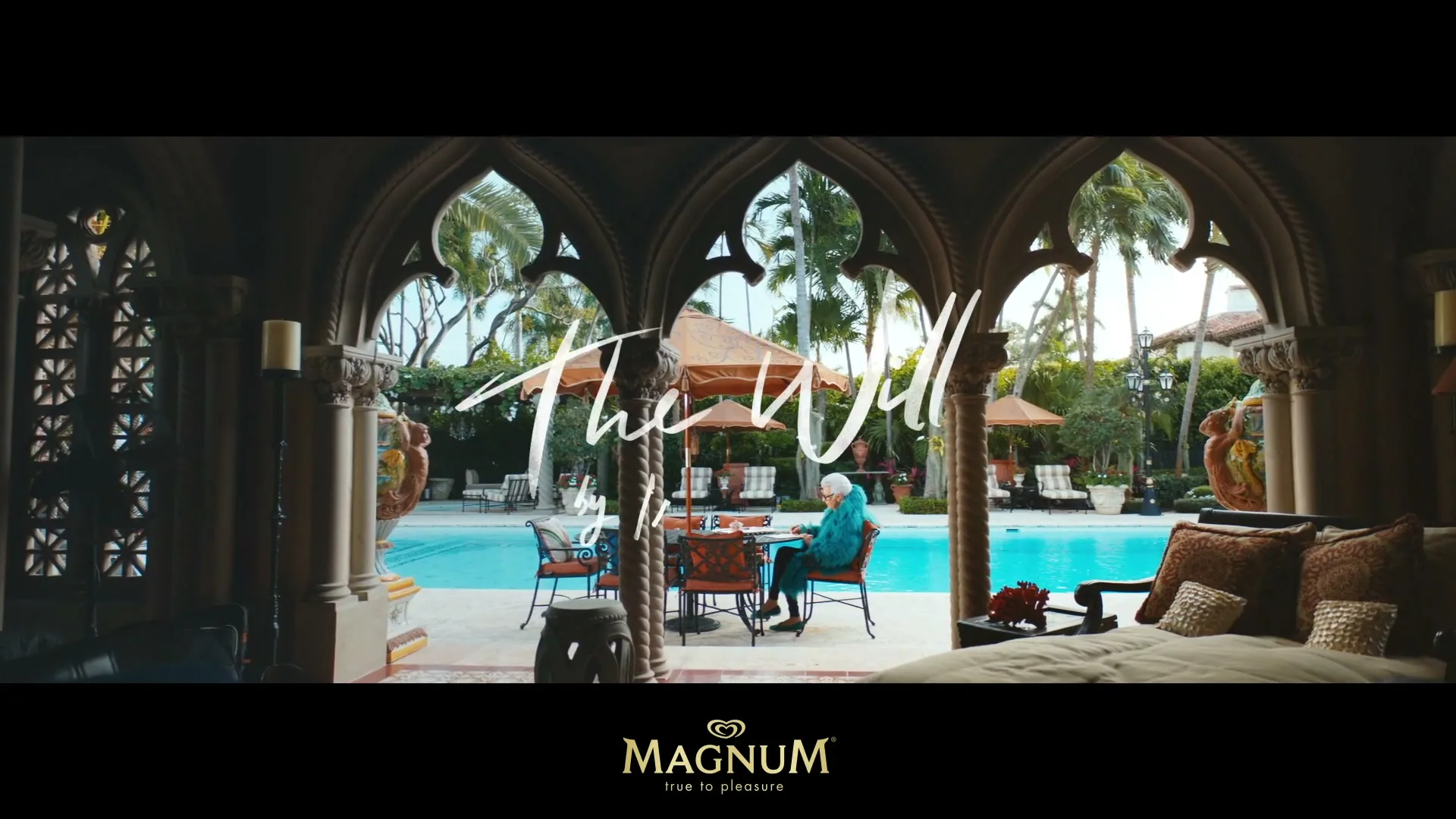 An older woman in a blue outfit sits at a table by a pool, seen through ornate archways of a luxurious interior.