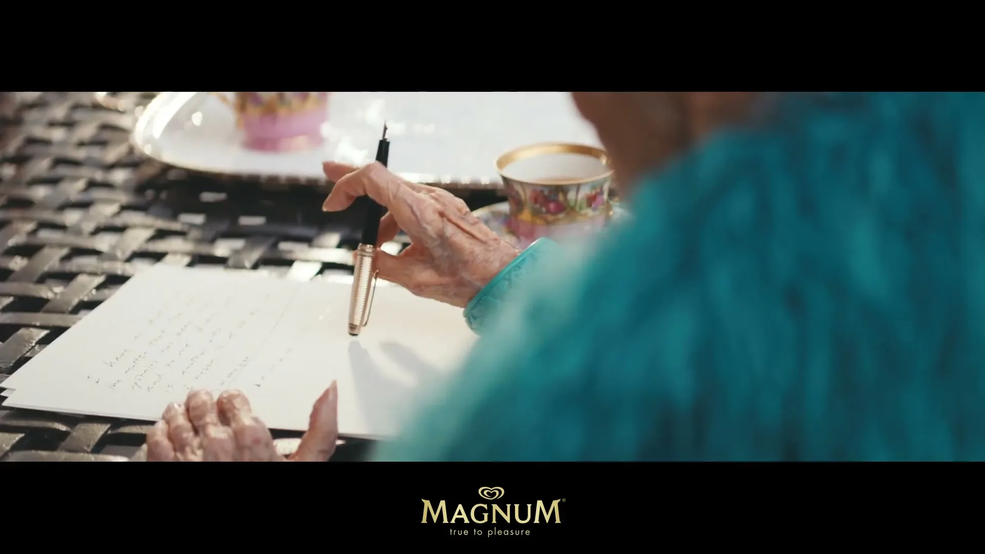 An older person with wrinkled hands, wearing a teal top, writes a letter with a pen at a table outdoors, with a teacup in the background.