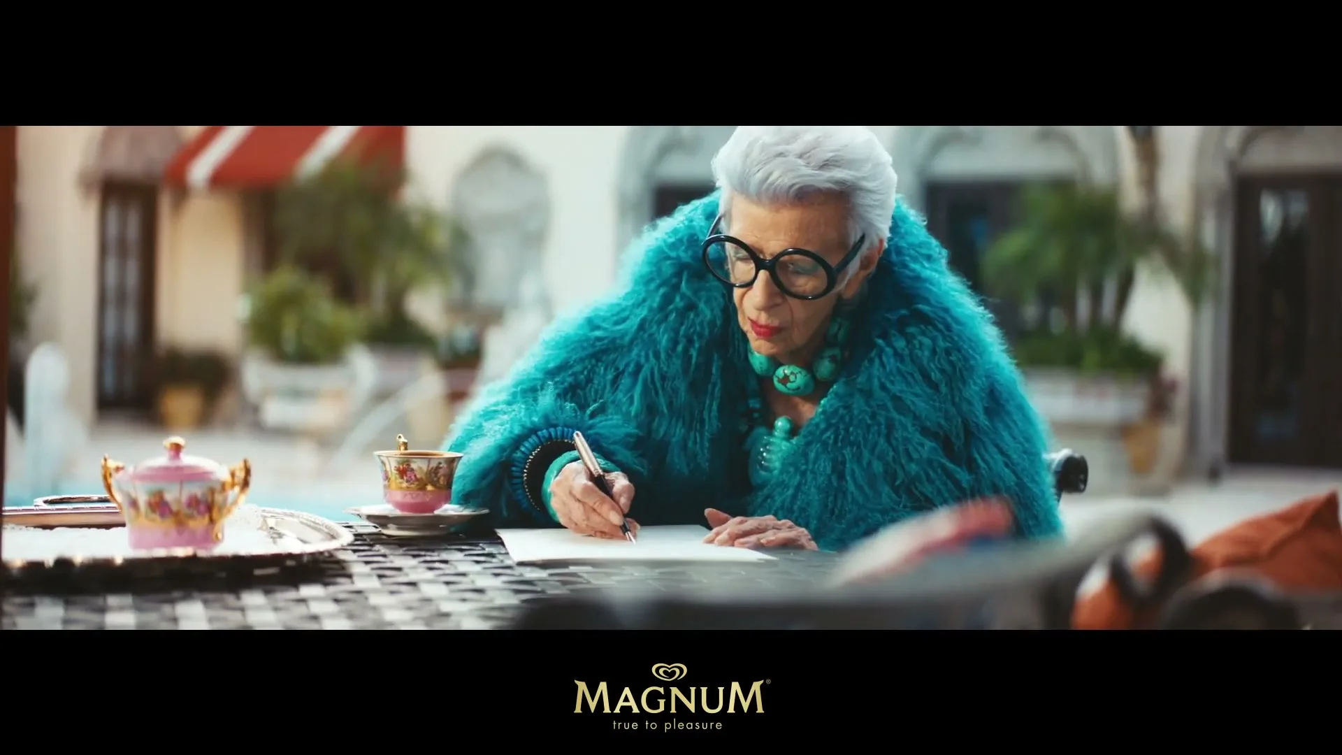 An older woman with white hair and large glasses, wearing a vibrant teal fur coat, writes at an outdoor table in a luxurious setting.