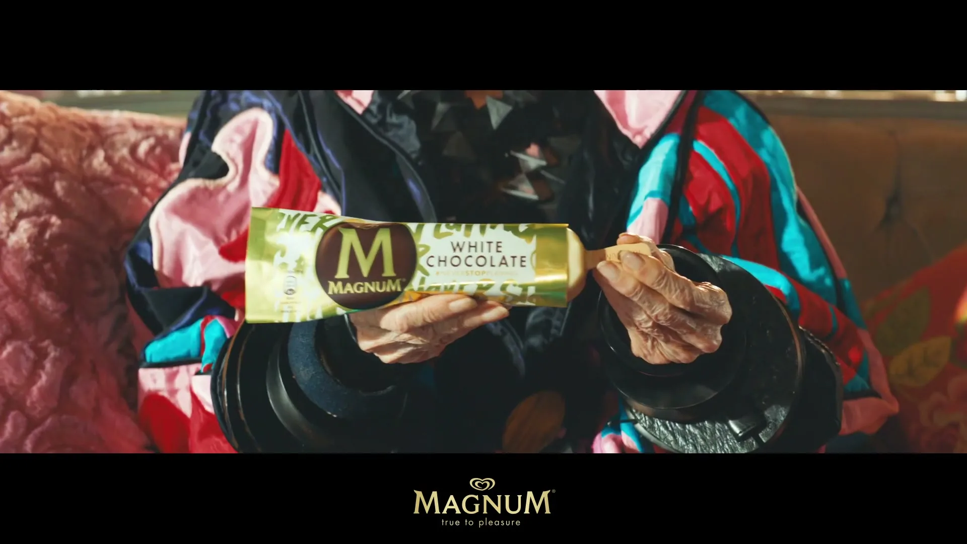 An older person with wrinkled hands, wearing a colorful jacket, holds a Magnum White Chocolate ice cream bar, unwrapping it in an interior setting.