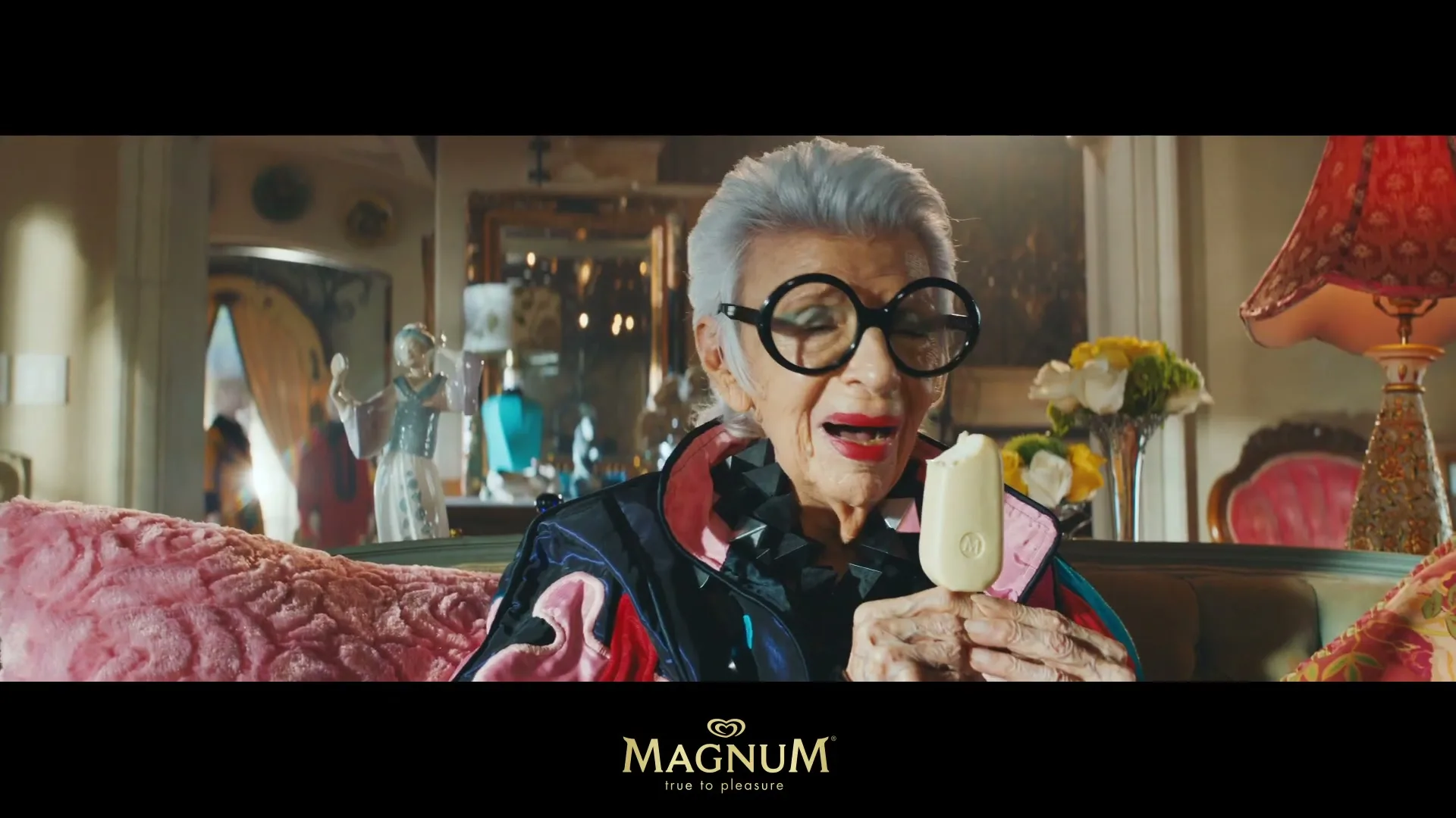 Iris Apfel, an elderly woman with grey hair and large glasses, sits on a couch, taking a bite of a white Magnum ice cream bar in a lavish interior.