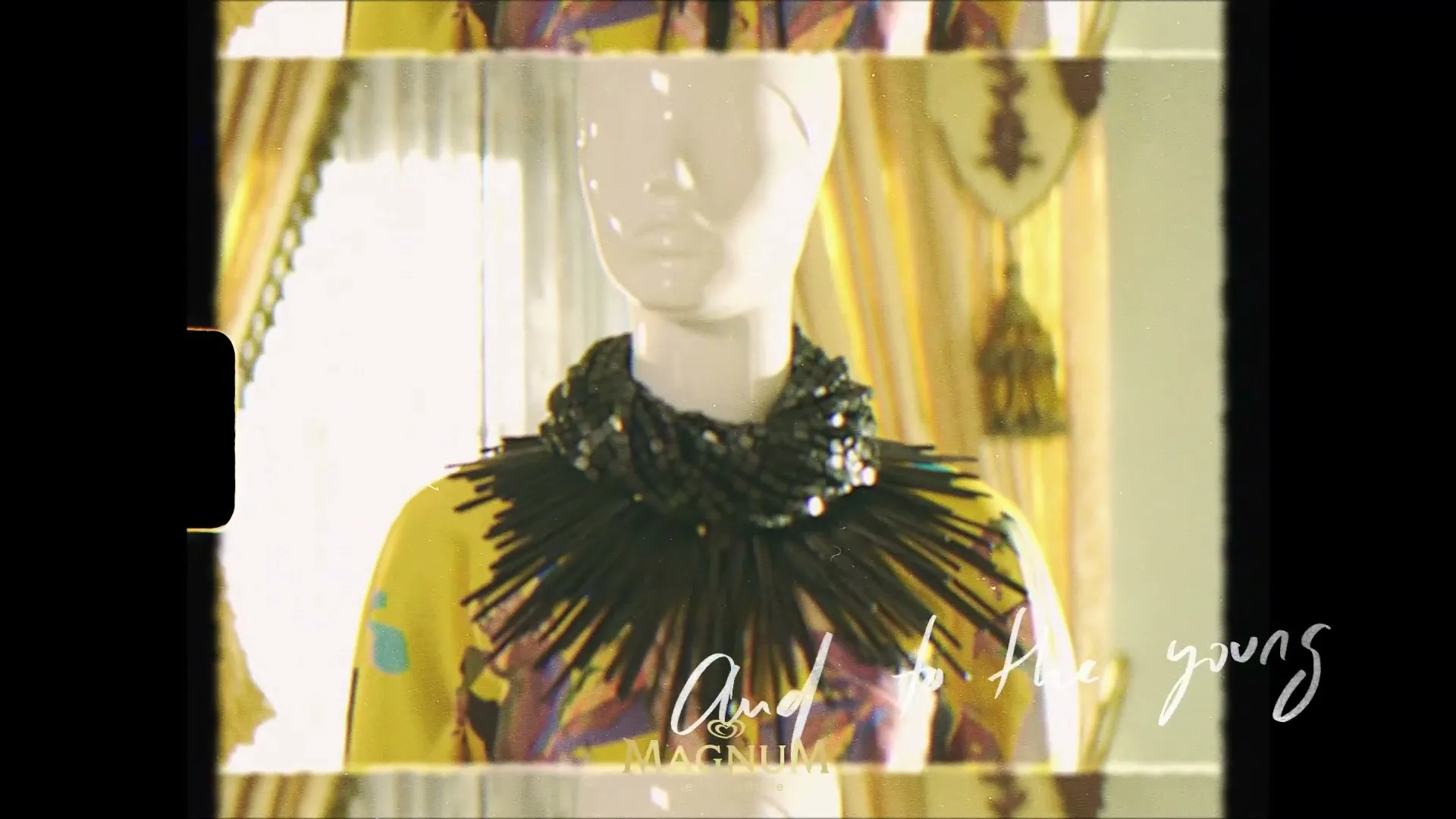 A white mannequin wears a yellow and purple patterned top with a black spiky necklace in a brightly lit room with yellow curtains.