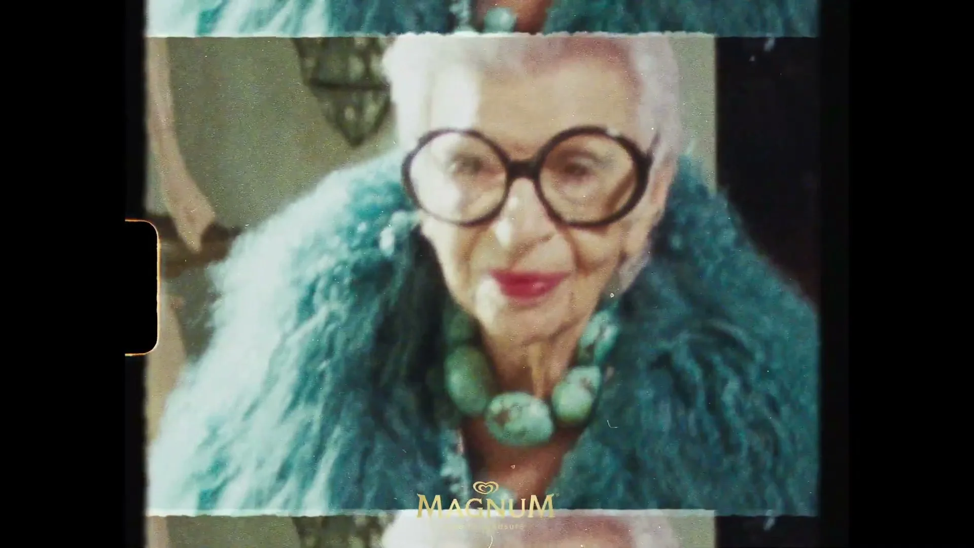 Iris Apfel, an elderly woman with white hair, large glasses, and a turquoise fur coat, smiles at the camera in an interior setting.