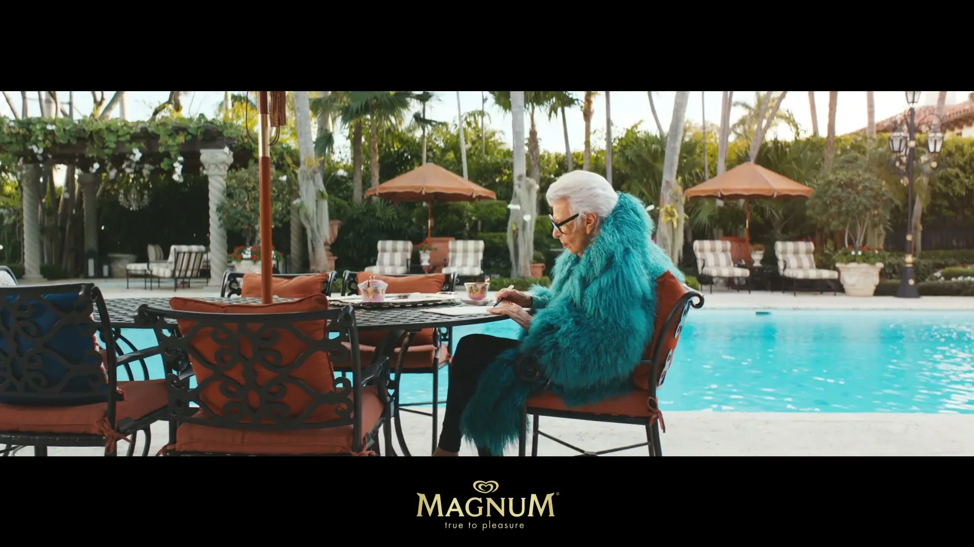 An elderly woman with white hair and a vibrant blue fur coat sits at a table by a swimming pool, looking down at something in her hands.