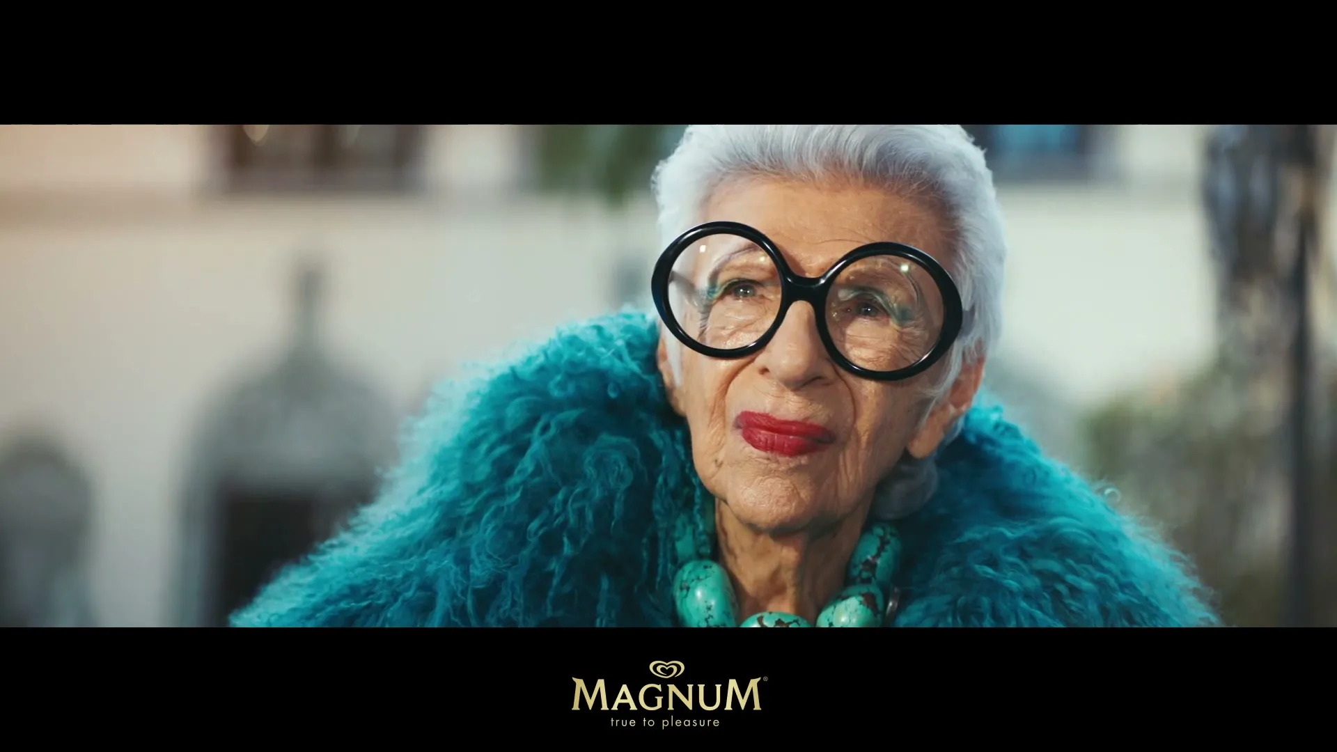 Iris Apfel, an elderly woman with white hair, large glasses, and a teal fur coat, looks up and to the right in an outdoor setting.
