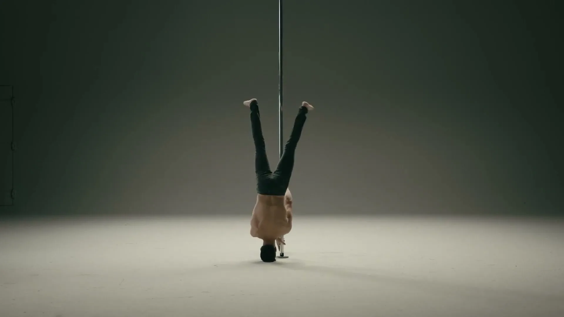 A shirtless man in dark pants performs a pole dance, doing a headstand and then a graceful pose, in a minimalist studio.